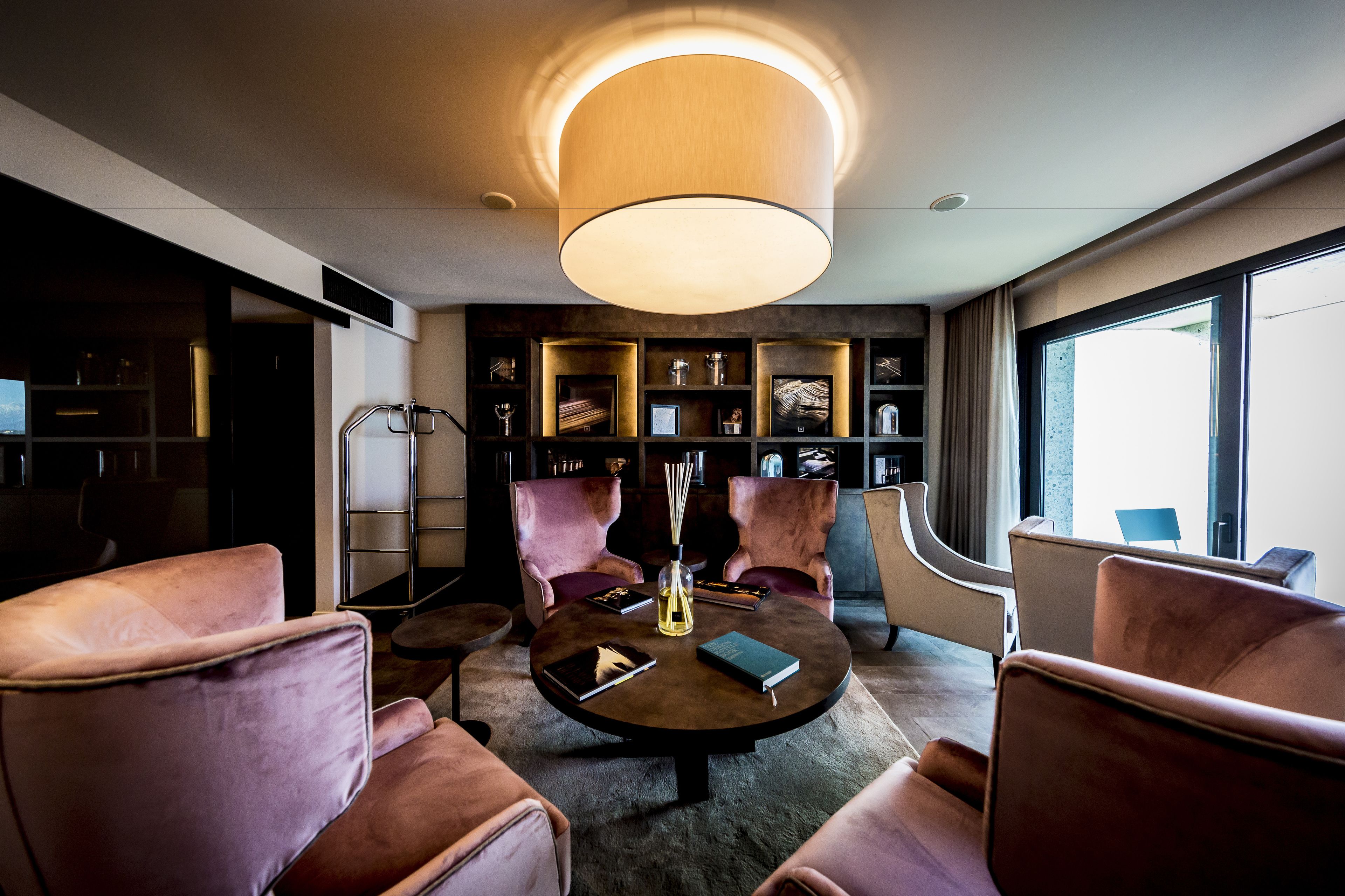 A modern living room with a round coffee table, two pink armchairs, a bookshelf, and a large window.