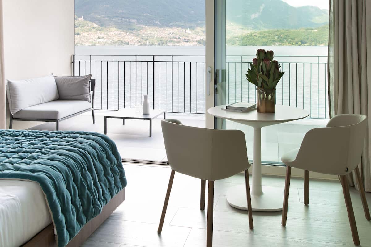 Een slaapkamer met een bed, een balkon, een tafel met stoelen en uitzicht op een meer en bergen.