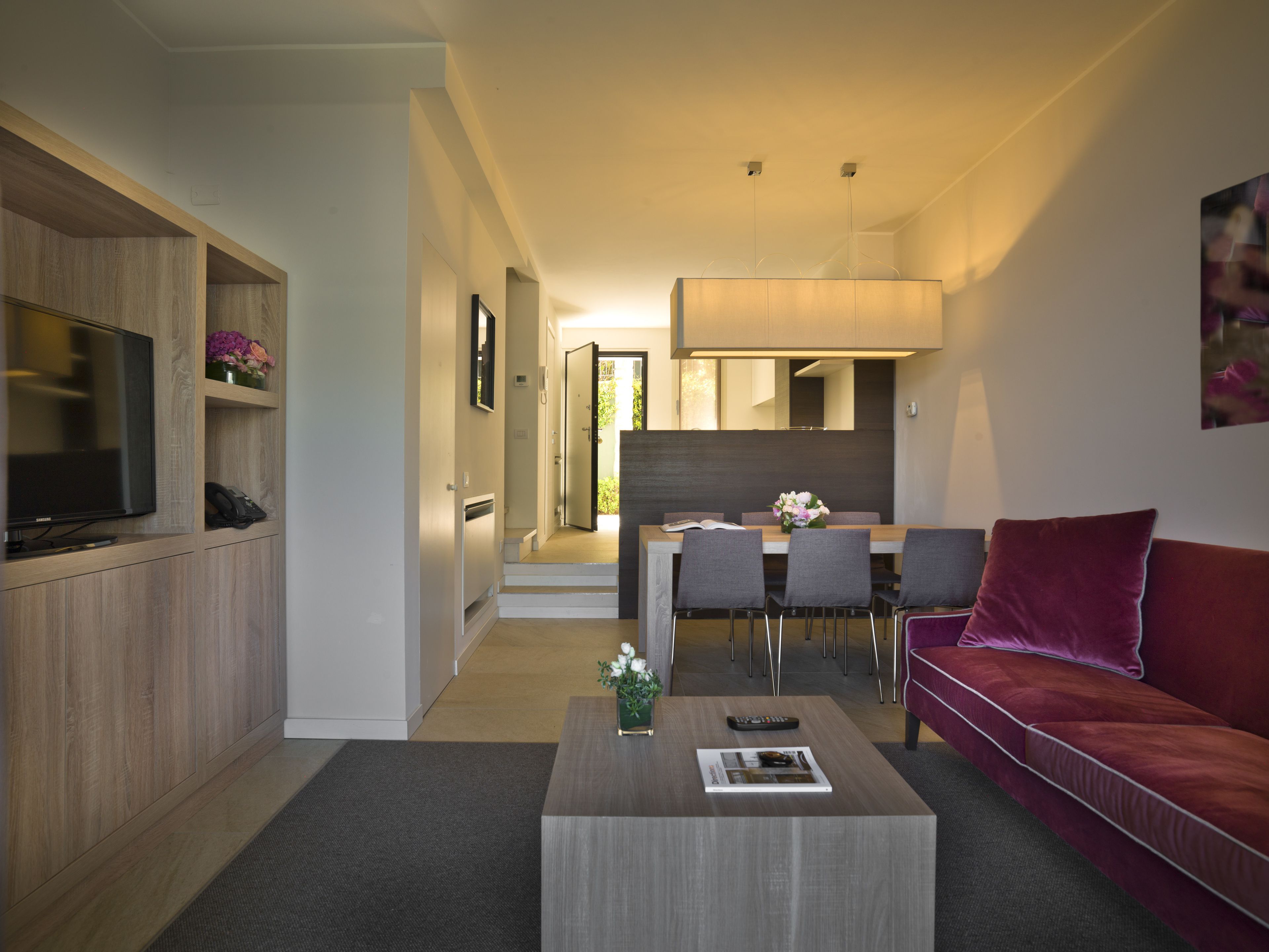 A modern living room with a flat-screen TV, a coffee table with a magazine, a red sofa, and a dining area with chairs and a table.