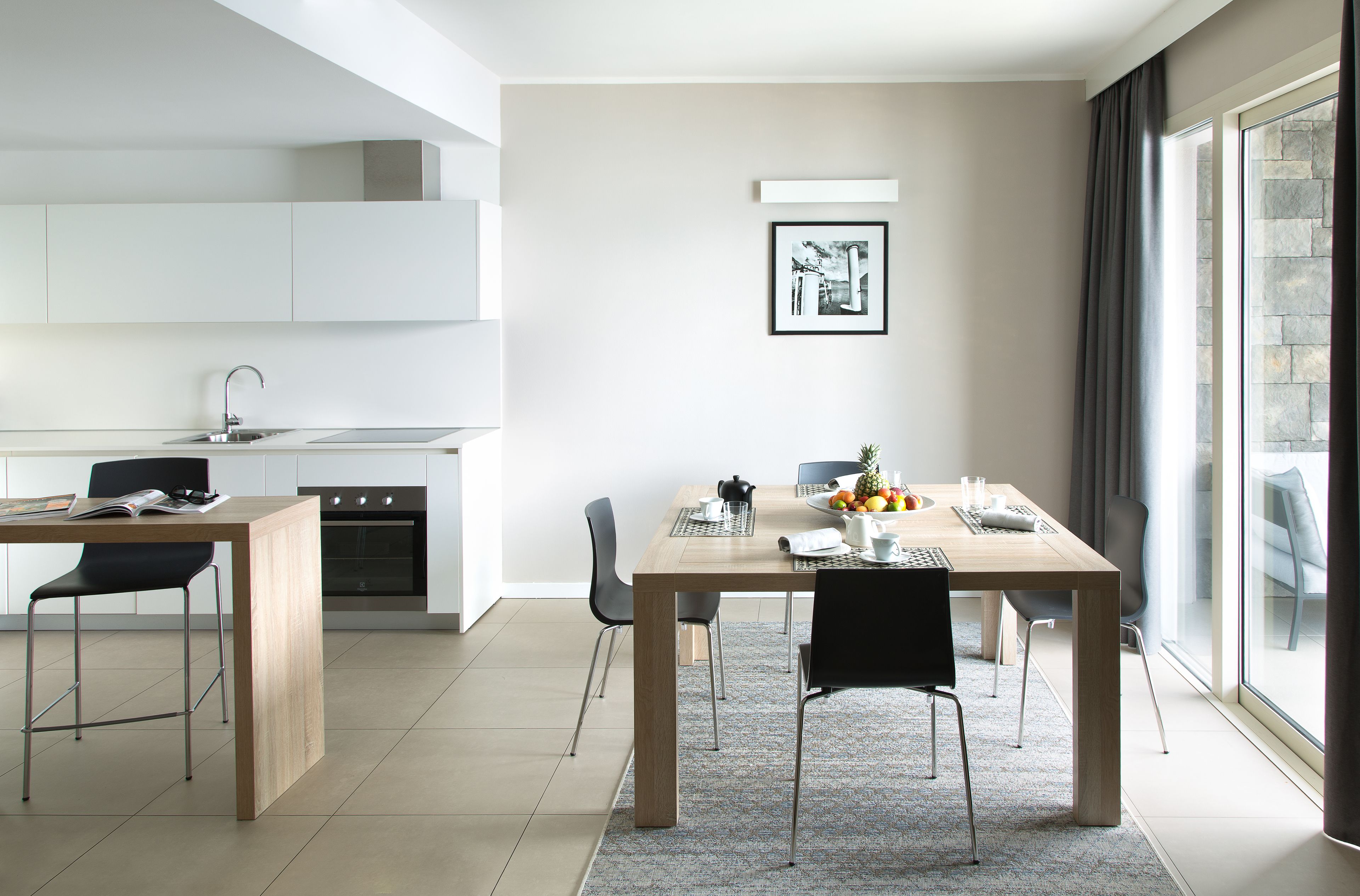 A modern kitchen with white cabinets, a dining area with a wooden table and chairs, and a large window with curtains.