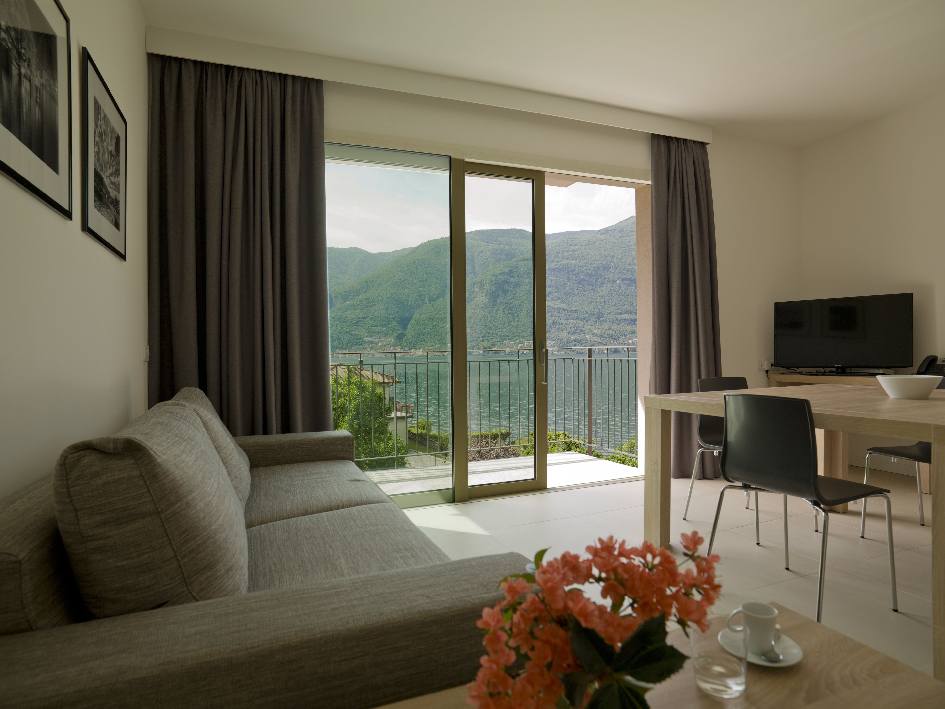 A modern living room with a sofa, a dining table with chairs, a TV, and a view of a lake and mountains through large windows.