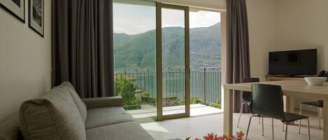 A modern living room with a sofa, a dining table with chairs, a TV, and a view of a lake and mountains through large windows.