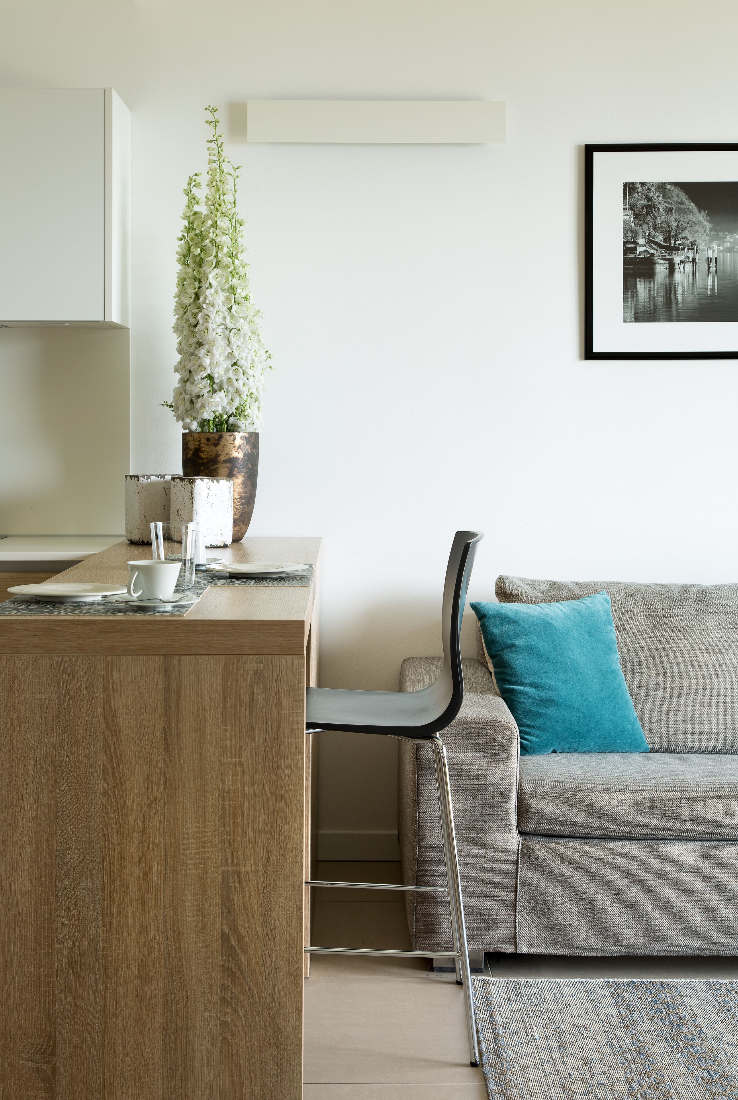 Un salon moderne avec une table basse en bois, un canapé gris agrémenté d’un coussin turquoise, un grand arrangement floral blanc et une photographie en noir et blanc encadrée accrochée au mur.