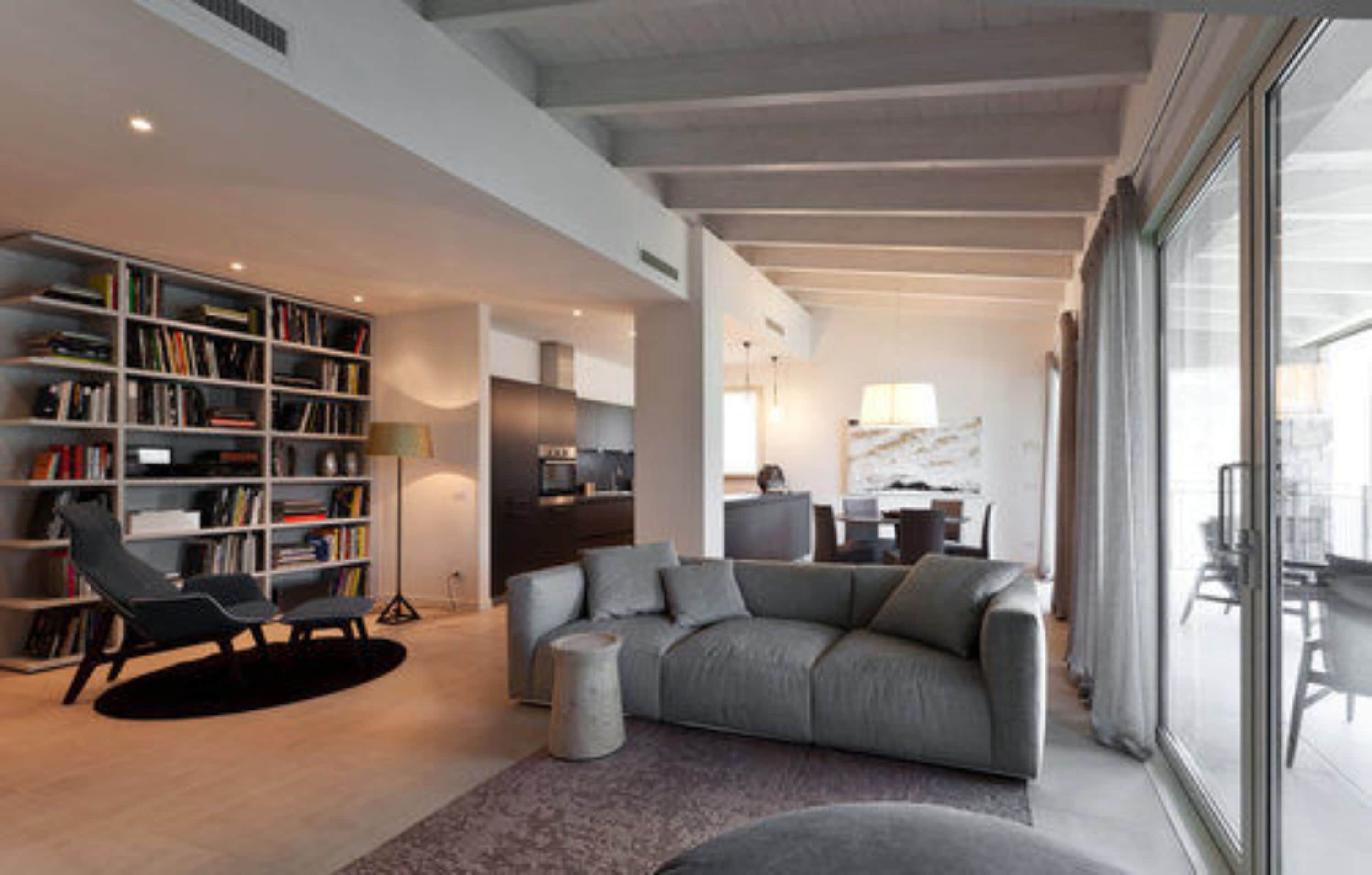A modern living room with a grey sofa, a bookshelf, and a dining area with a table and chairs.