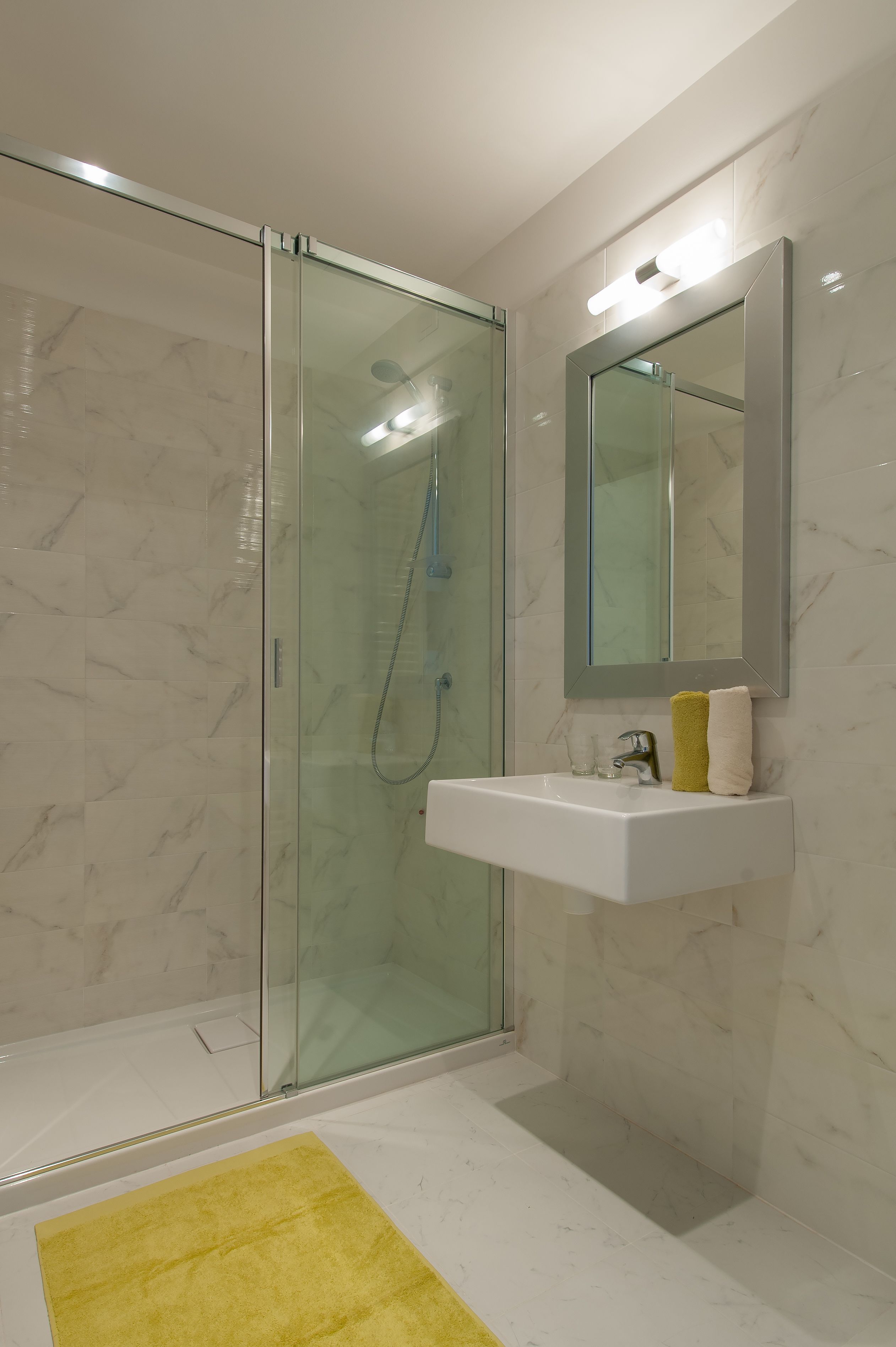 A modern bathroom with a glass shower enclosure, a white sink with a mirror above it, and a yellow bath mat on the floor.