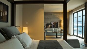 A modern hotel room with a large bed, a sofa, a dining table, and a view of the sea through large windows.