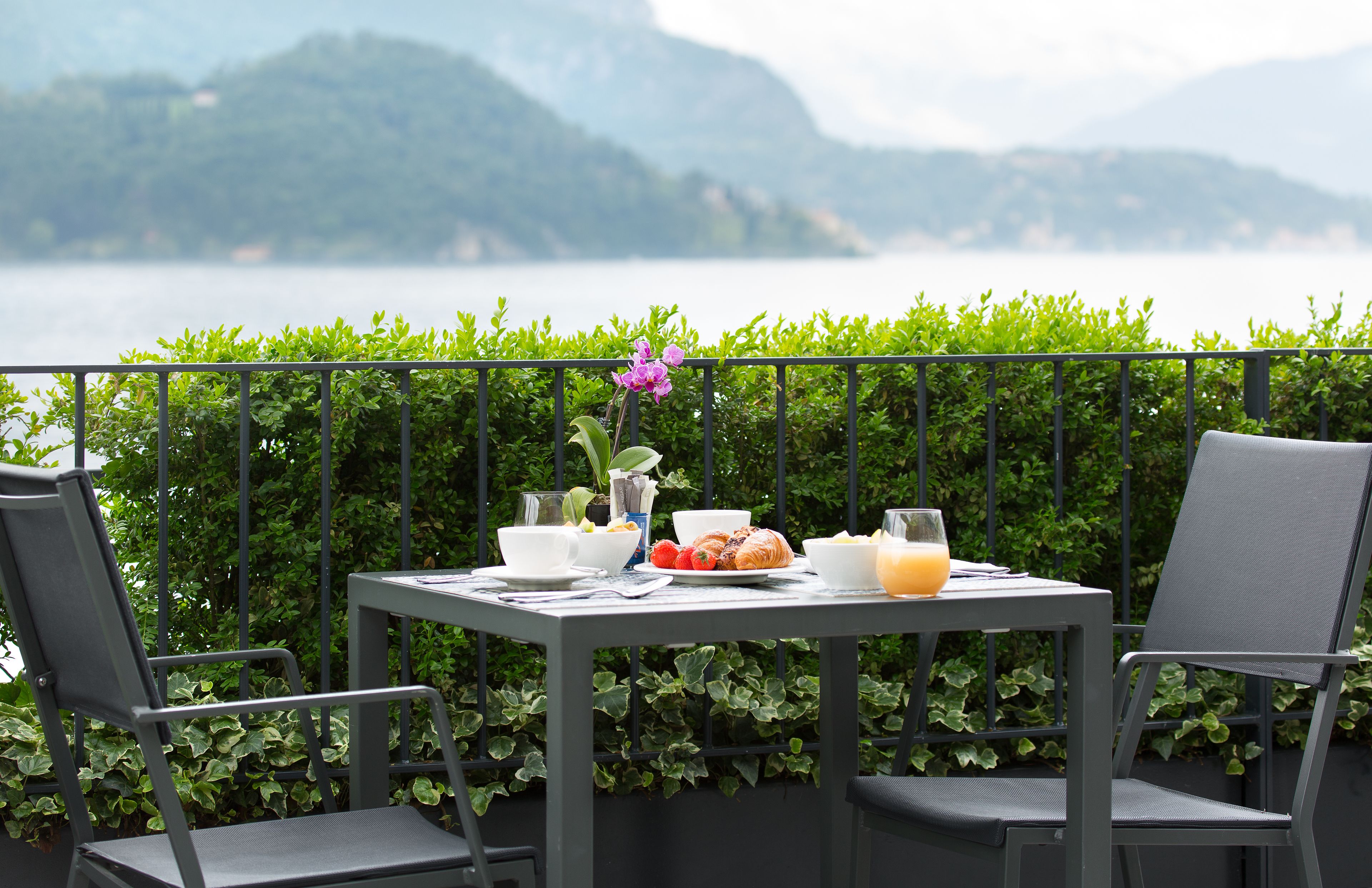 Une table dressée avec des aliments pour le petit-déjeuner, notamment des croissants, des fruits et du jus, sur un balcon offrant une vue sur un lac et des montagnes.