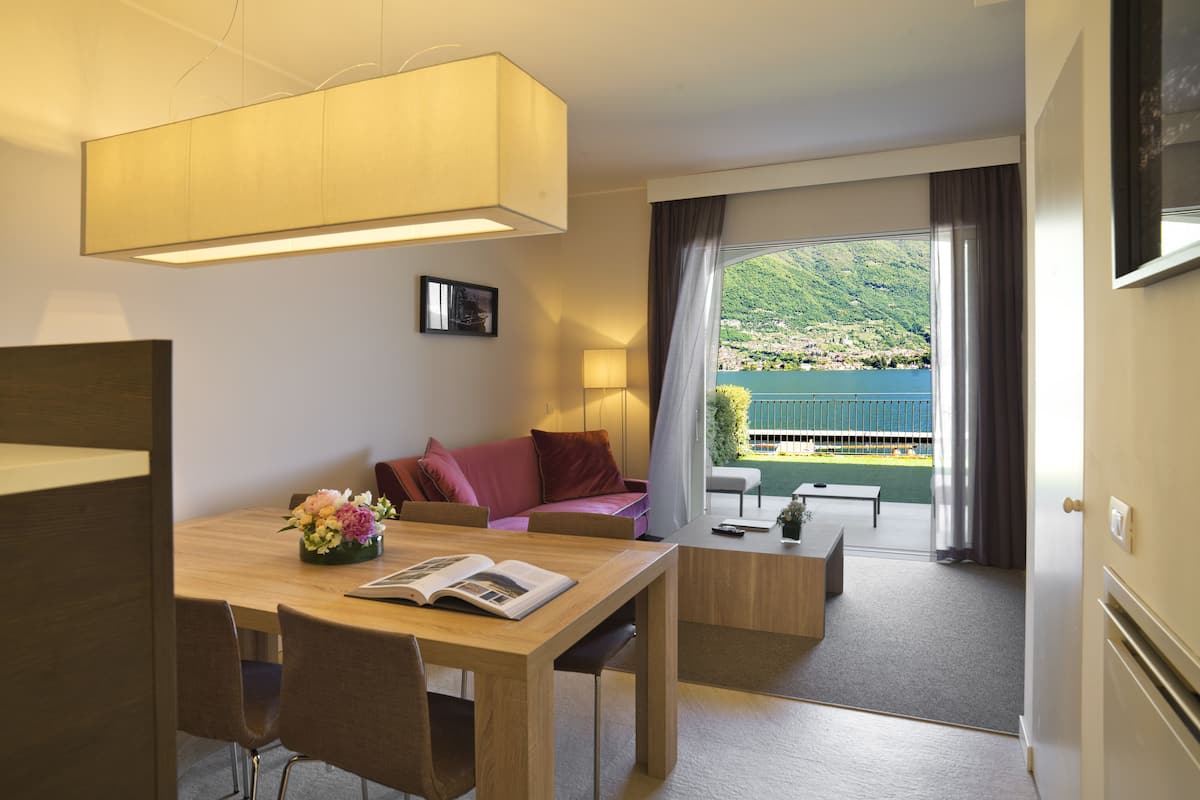 Een moderne hotelkamer met een zithoek, een sofa, een balkon met uitzicht op een meer en heuvels, en een groot raam.