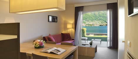 A modern hotel room with a dining area, a sofa, a balcony with a view of a lake and hills, and a large window.