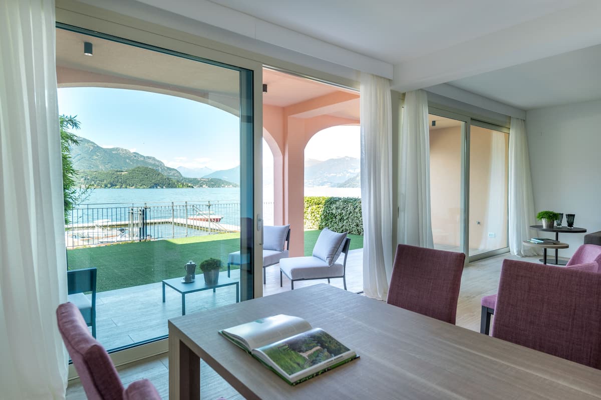 Een moderne kamer met een grote schuifglazen deur die uitkomt op een balkon met uitzicht op een meer en bergen. Er staat een tafel met een boek erop en enkele stoelen.