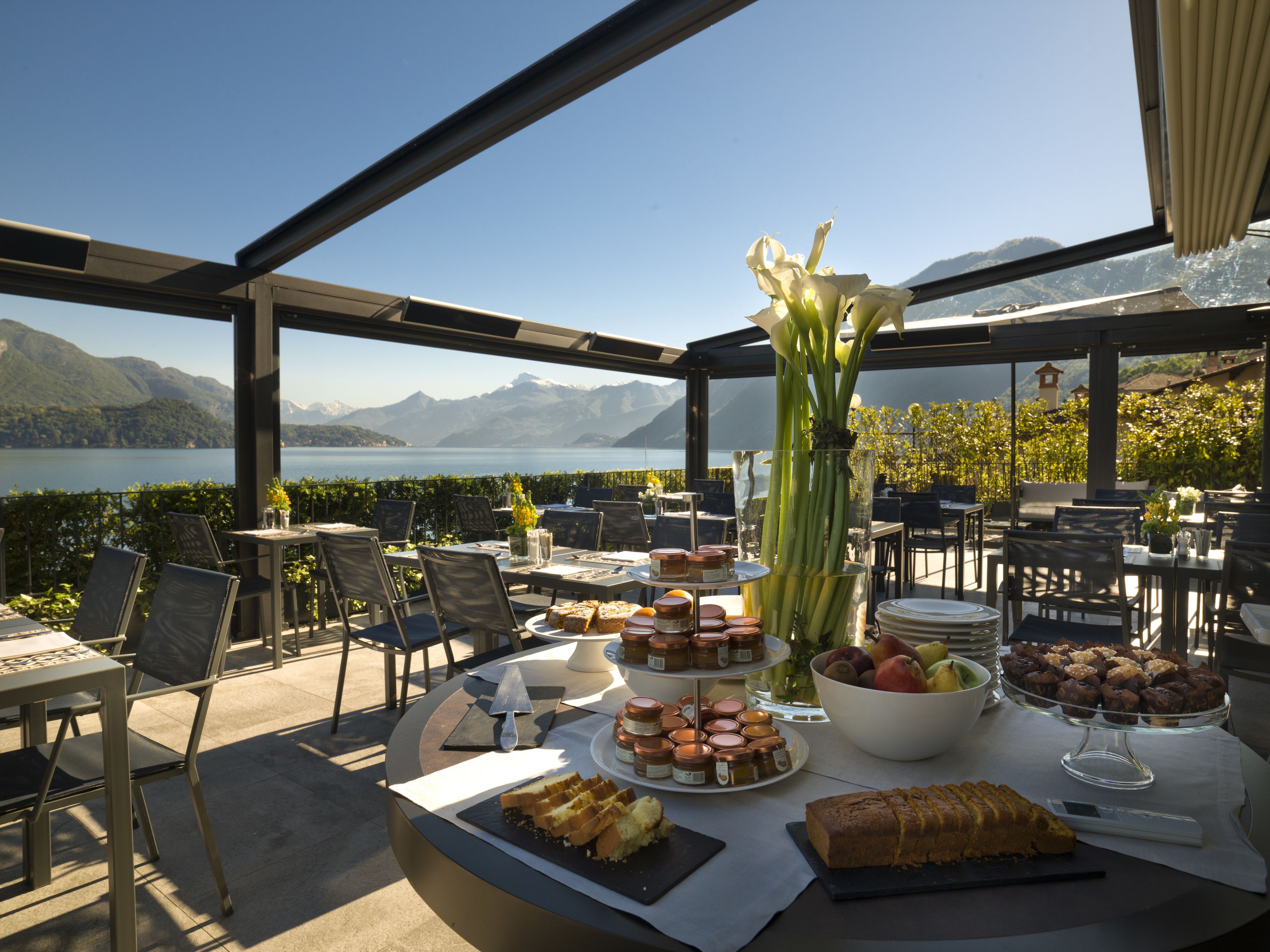 An outdoor dining area with tables and chairs, a variety of food and drinks, and a view of a lake and mountains.