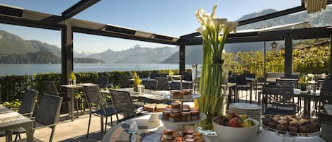 An outdoor dining area with tables and chairs, a variety of food and drinks, and a view of a lake and mountains.