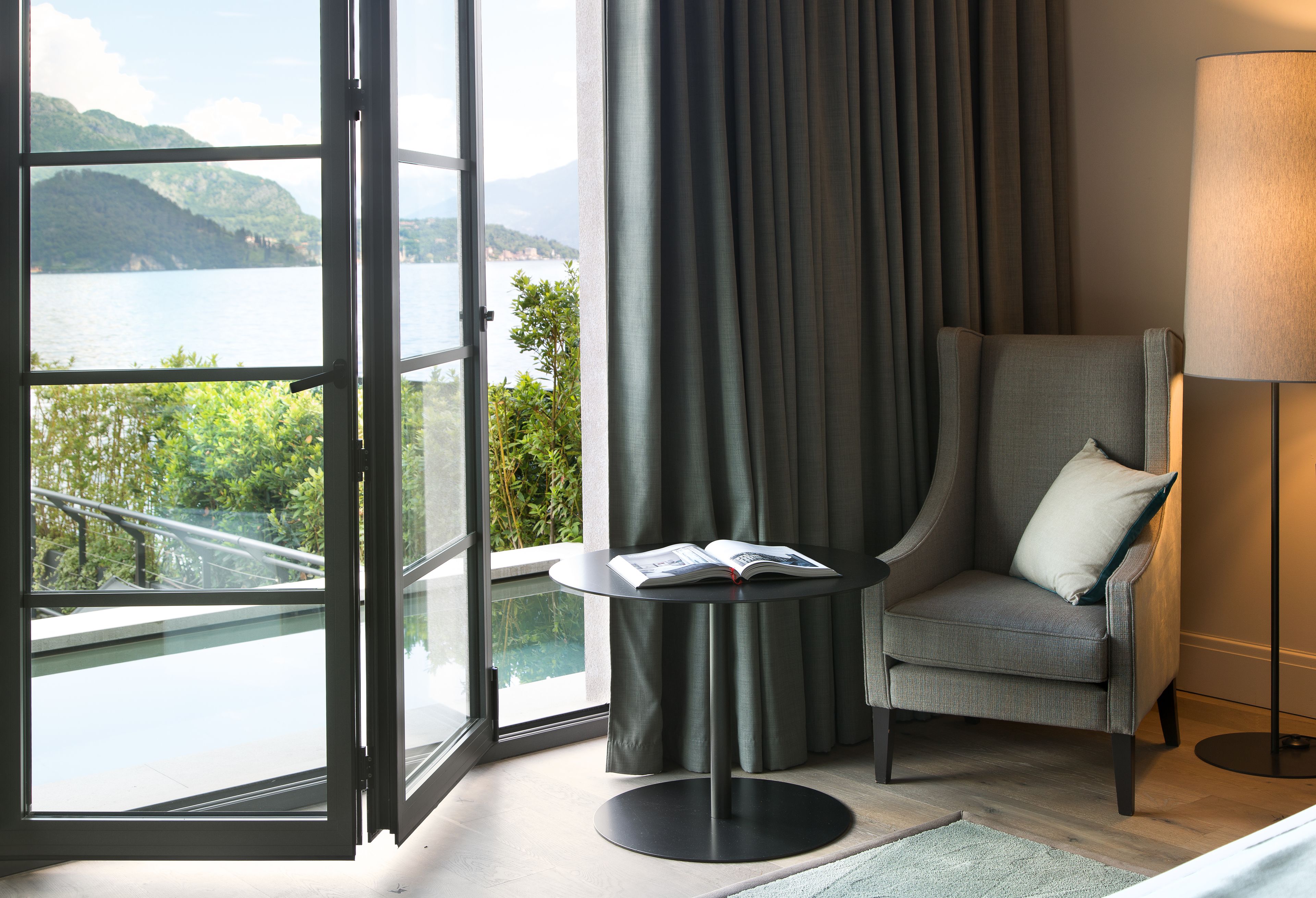 A room with a view of a lake, a chair with a cushion, a table with a book, and a lamp.