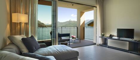A modern hotel room with a large bed, a sofa, a balcony with a view of the lake, and a TV on a stand.