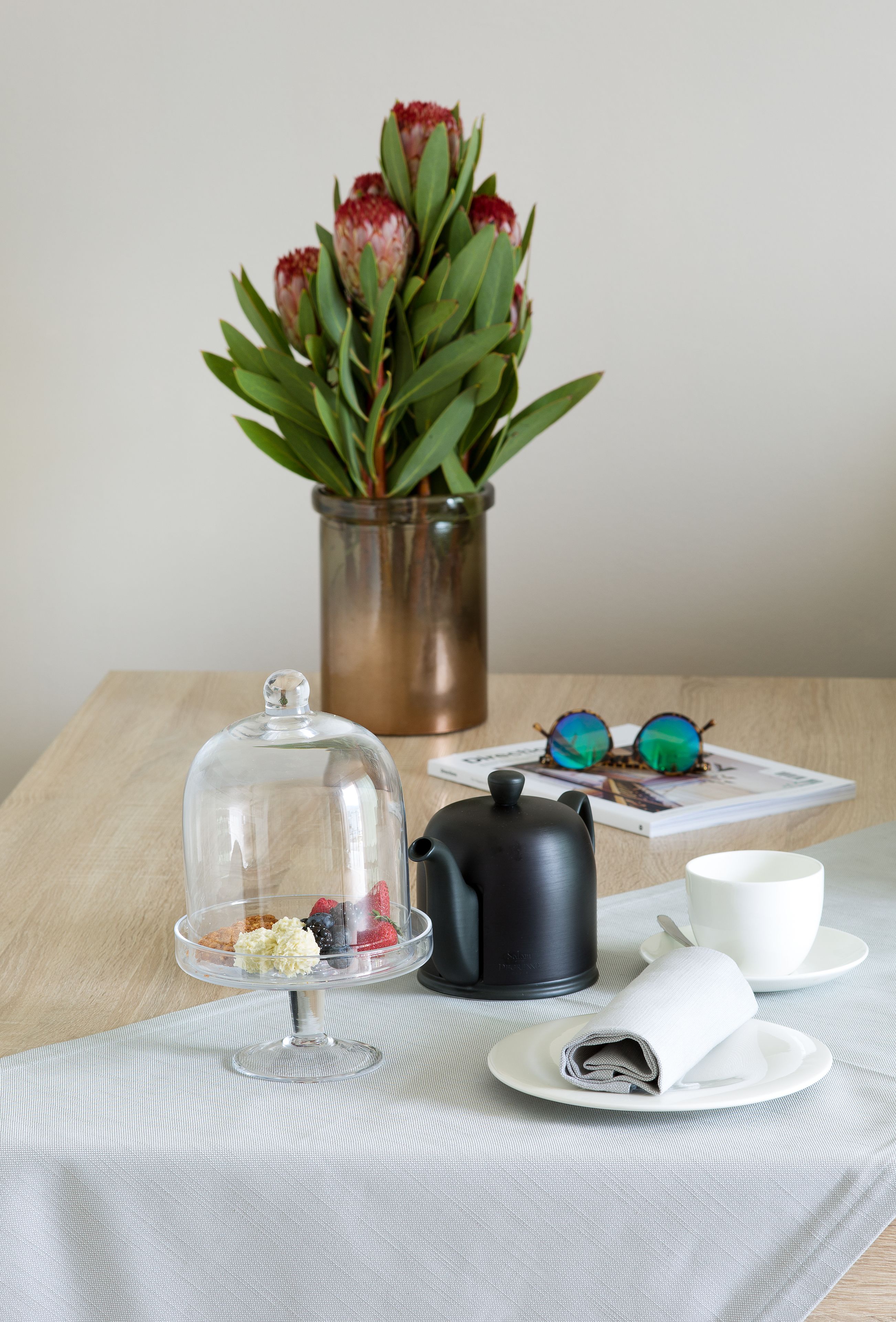 Une table avec un cloche en verre, une théière noire, une tasse blanche, une serviette roulée, une assiette à dessert, une paire de lunettes de soleil et un vase avec des fleurs rouges.