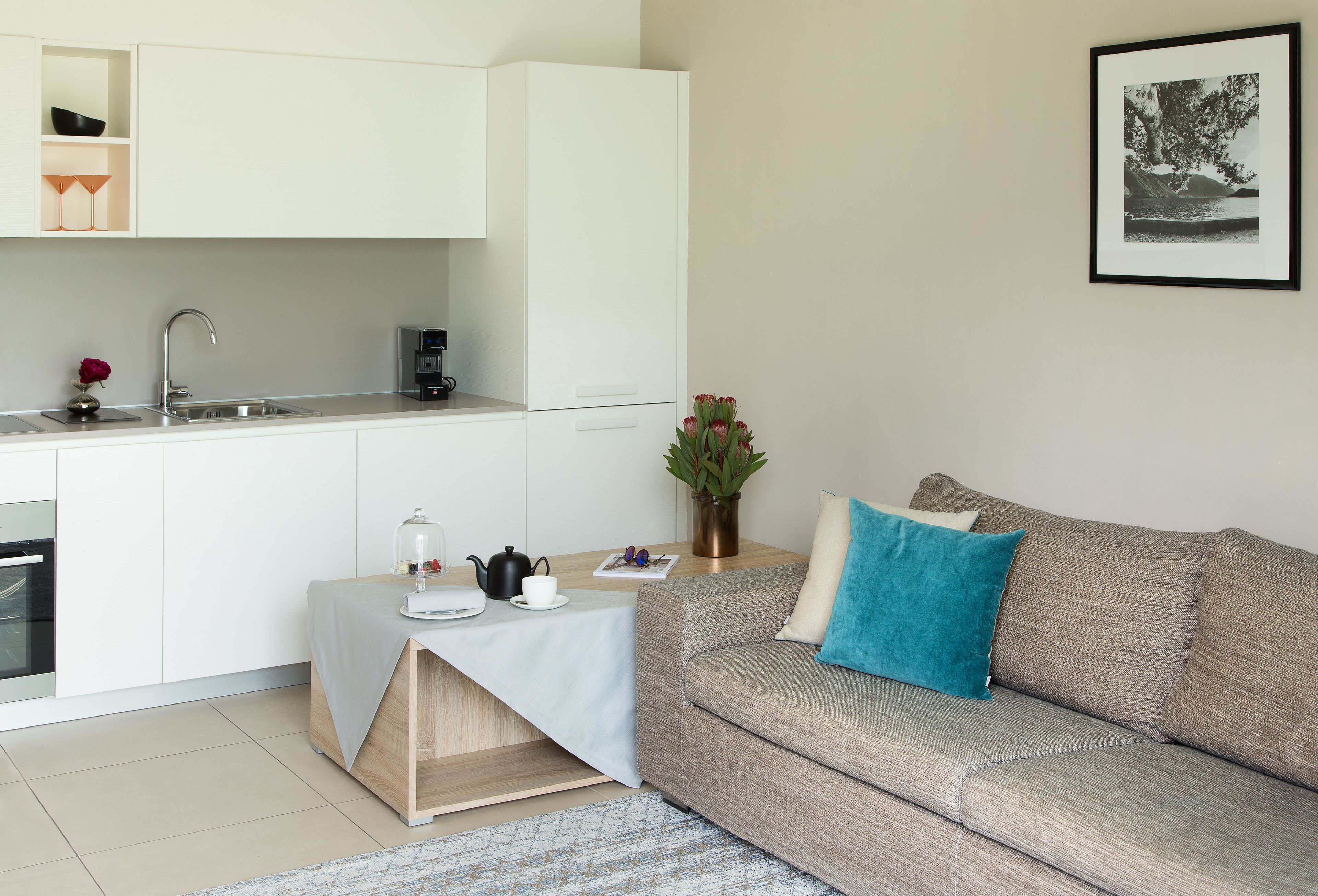 A modern kitchen with white cabinets, a sink, and a coffee machine. There is a dining area with a table, a vase with flowers, and a sofa with a blue pillow. A framed picture hangs on the wall.