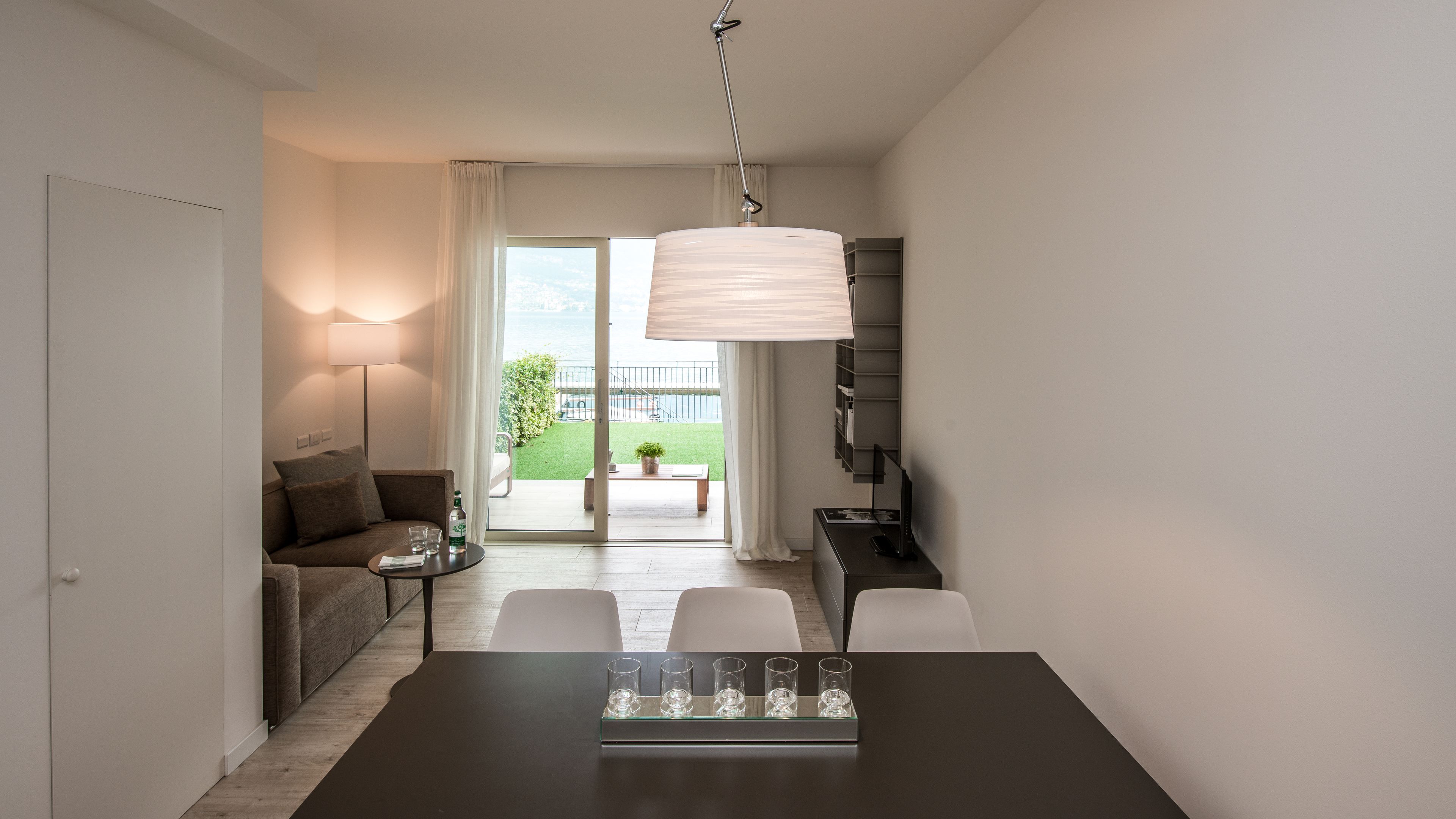 A modern hotel room with a dining area, a sofa, a table with glasses, and a view of a balcony with a garden.