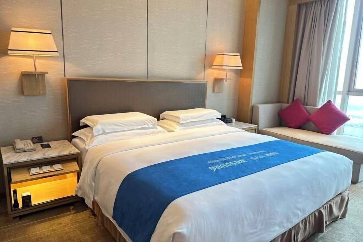Une chambre d’hôtel avec un lit, des lampes de chevet, une table de chevet, une fenêtre avec des rideaux et un banc avec des coussins.