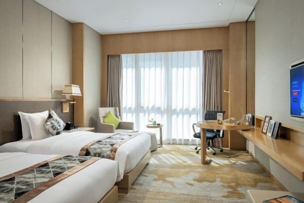 Habitación de hotel con dos camas, un escritorio, una silla, un televisor y un ventanal con cortinas.