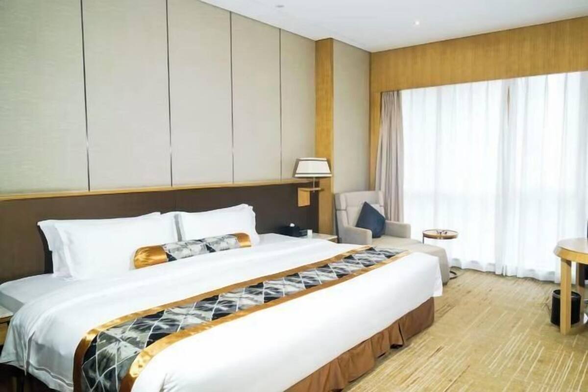 Une chambre d’hôtel avec un grand lit, une table de chevet, une chaise, une lampe et une vue sur l’extérieur par une fenêtre.