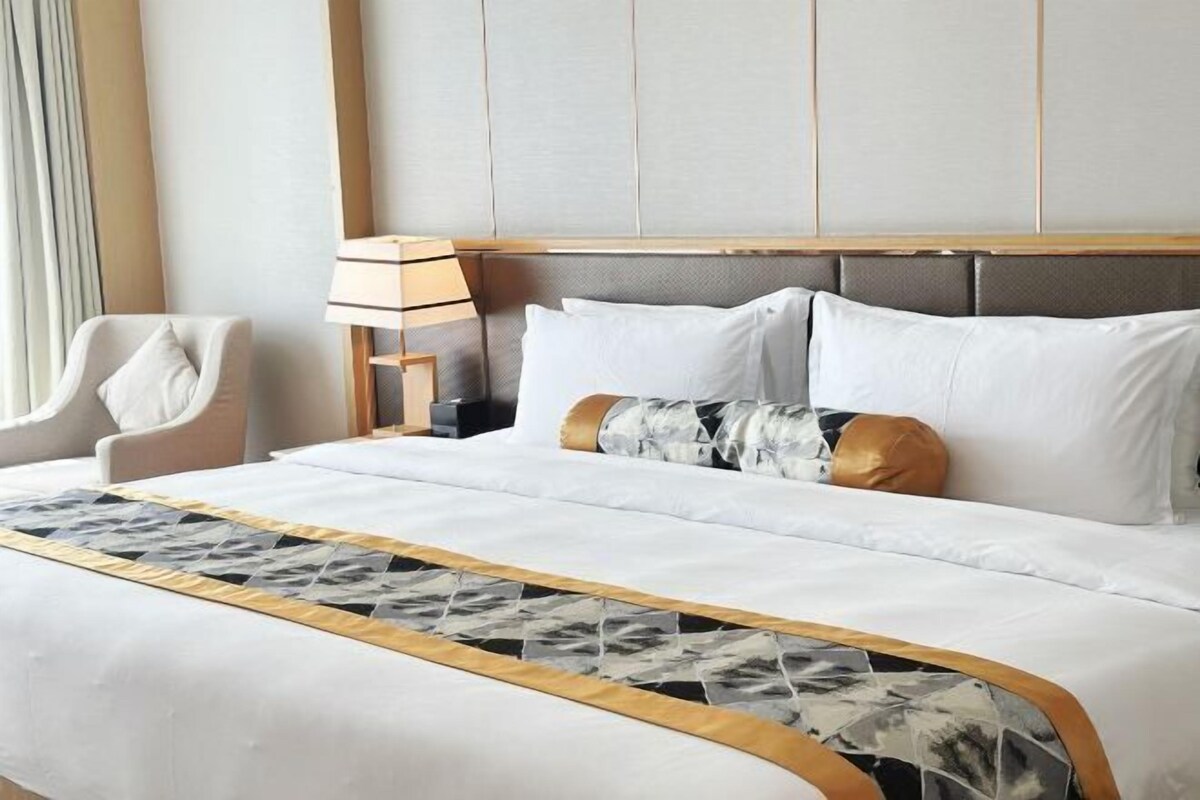 Una cama bien tendida con sábanas blancas y almohadas decorativas, una lámpara de noche y una silla.
