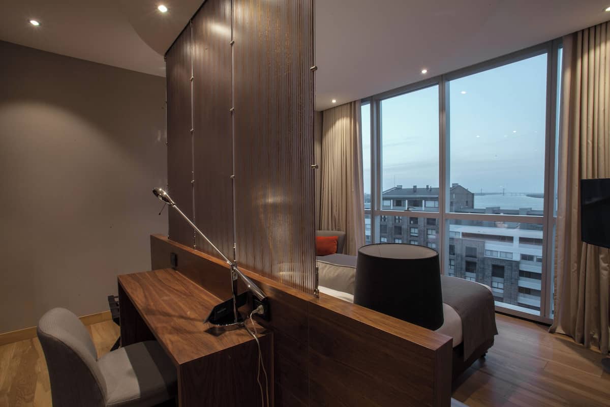 Ein modernes Hotelzimmer mit einem Holztisch, einer gemütlichen Couch, einem großen Fenster mit Blick auf die Stadt und einem Flachbildfernseher.