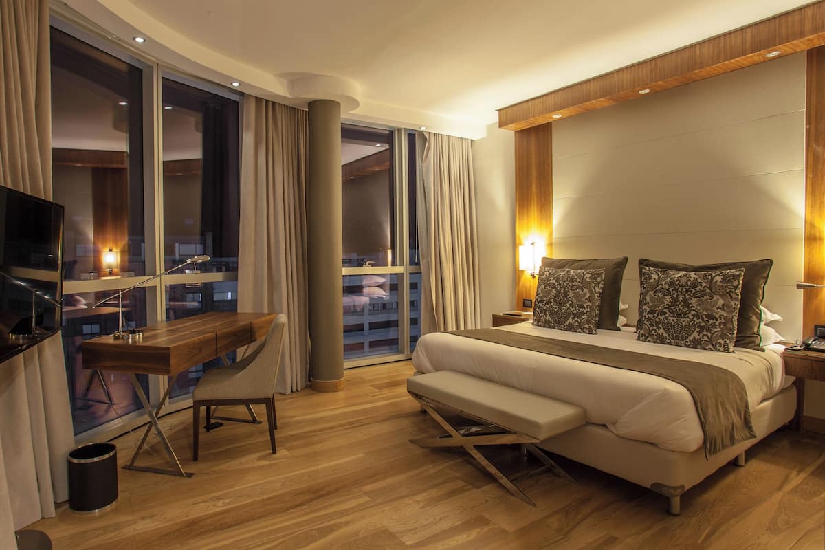 Ein Hotelzimmer mit Bett, Schreibtisch, Stuhl, Fernseher und Blick auf die Stadt durch große Fenster.