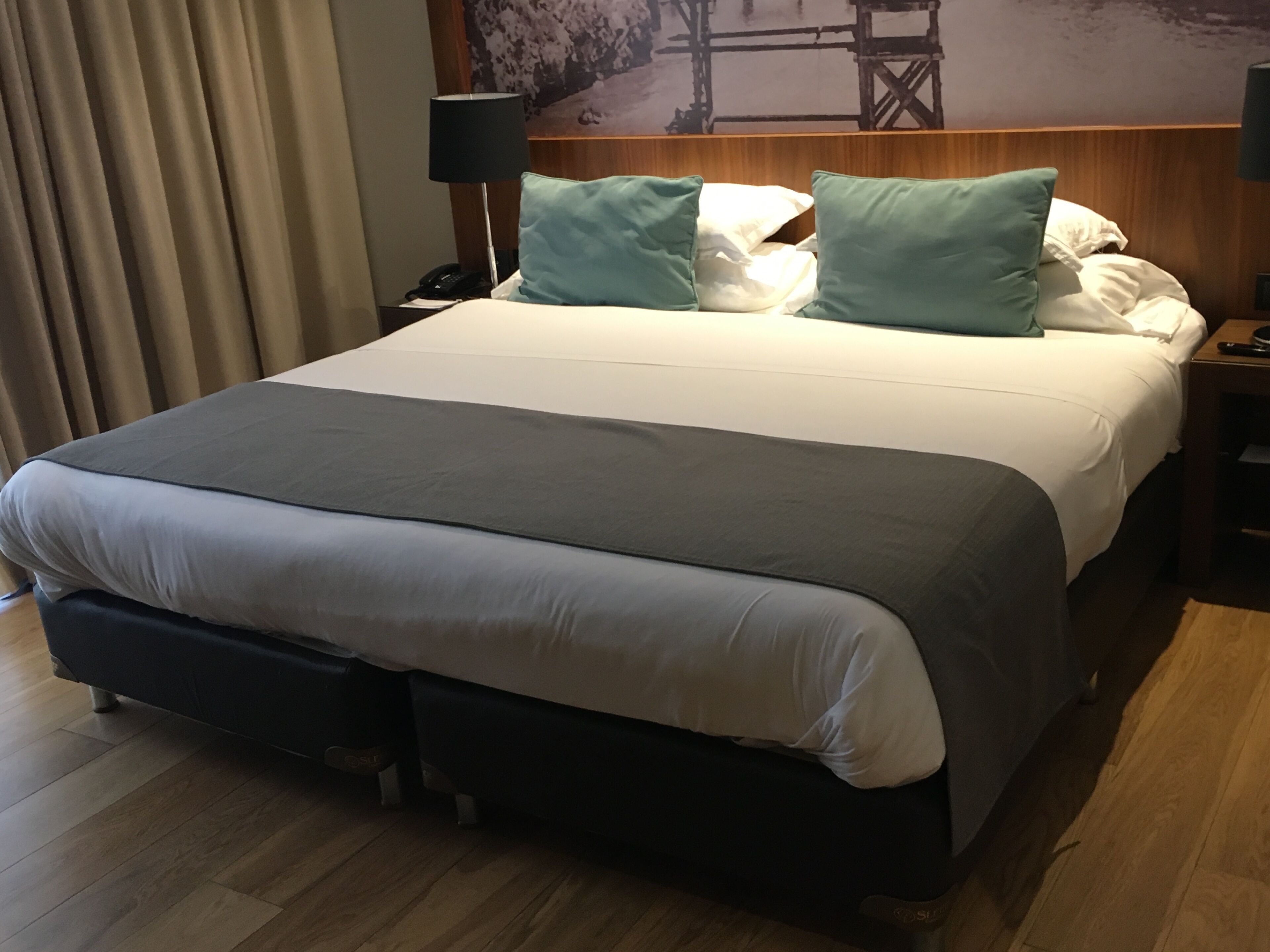 A hotel room with a large bed, a wooden headboard, a nightstand with a lamp, and a painting on the wall.
