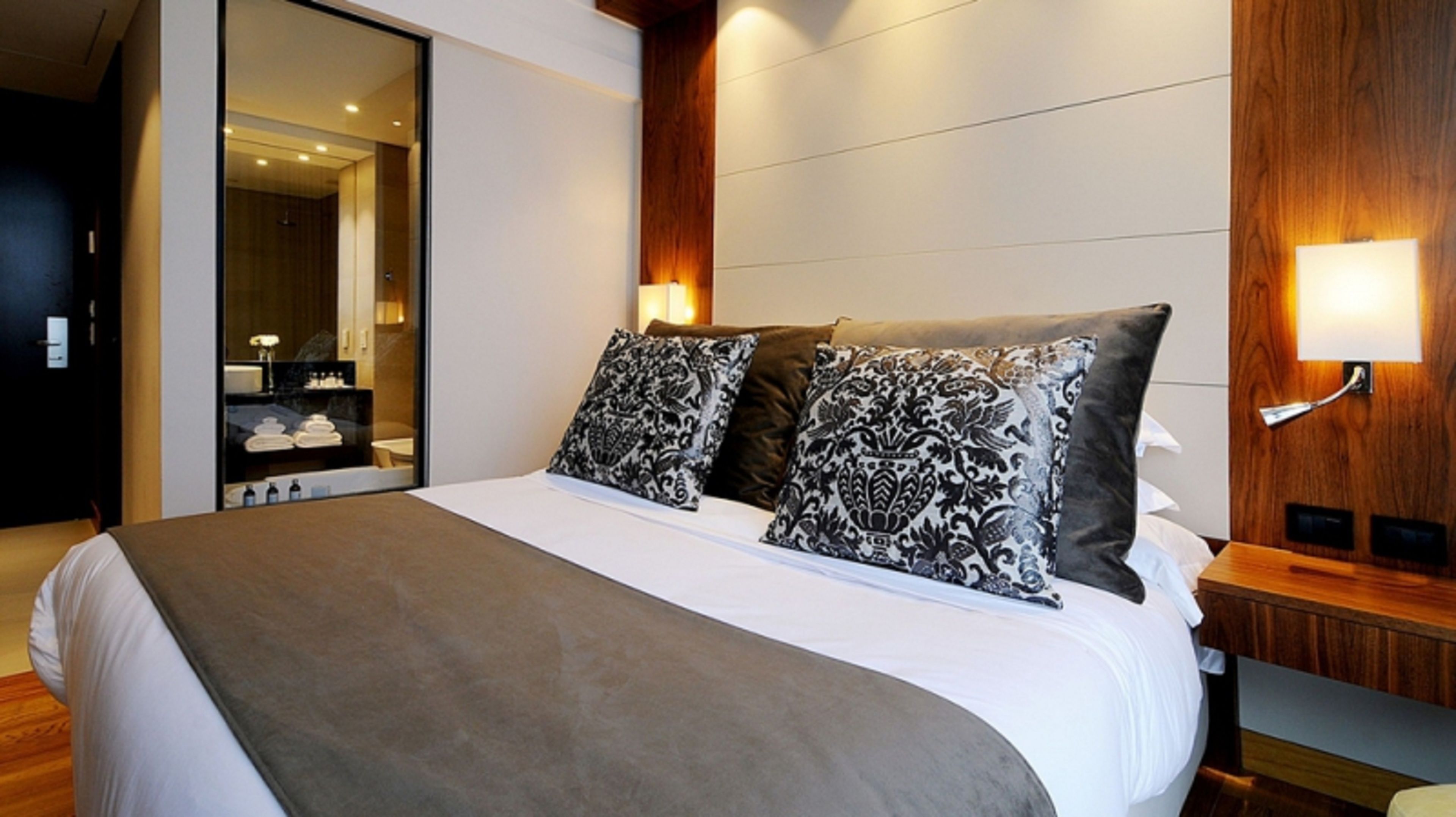 Une chambre d’hôtel moderne avec un grand lit, deux coussins à motifs, une tête de lit en bois, une table de chevet avec une lampe et une porte coulissante donnant sur la salle de bain.