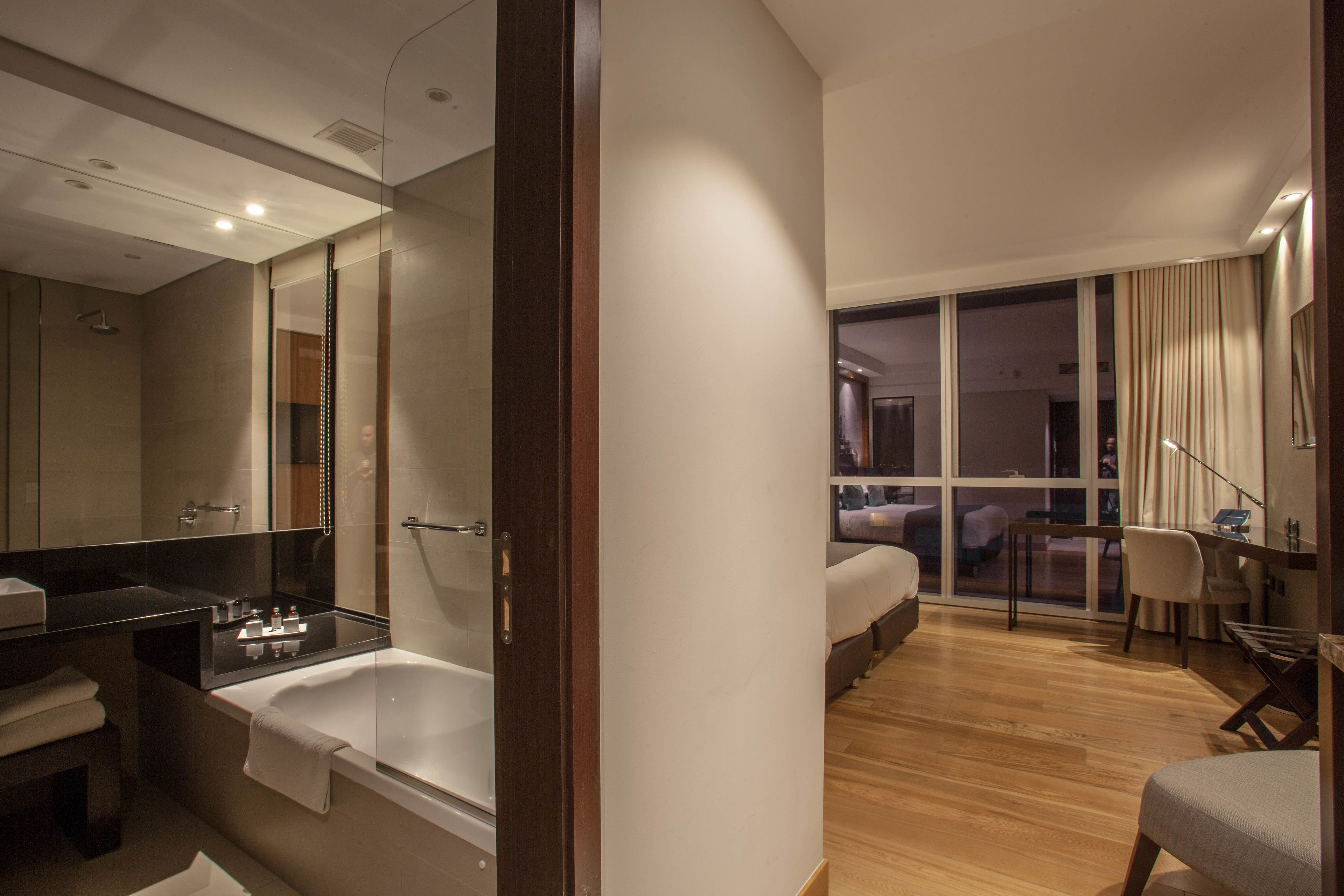 A modern hotel room with a large mirror, a bathtub, a desk, and a view of the city through large windows.