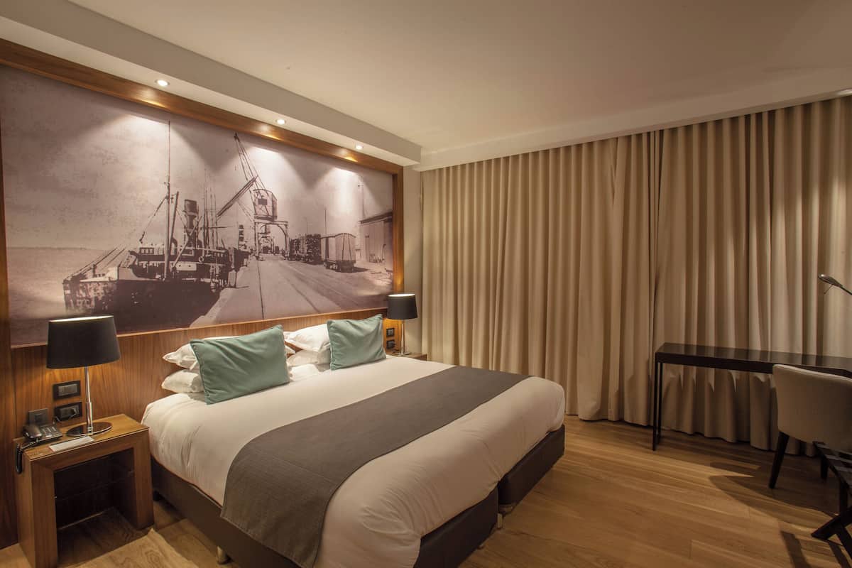 Ein Hotelzimmer mit einem großen Bett, einem Schreibtisch, einem Sessel, einem Nachttisch mit Lampe und einem großen Wandbild.