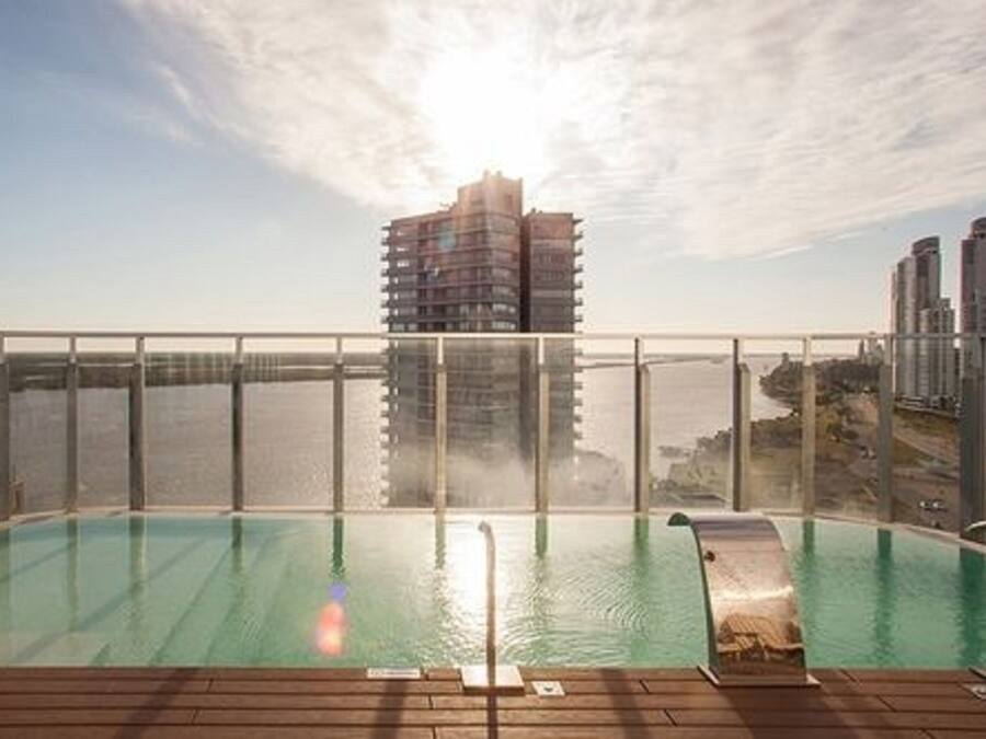 A rooftop pool with a glass barrier, overlooking a cityscape and a body of water.