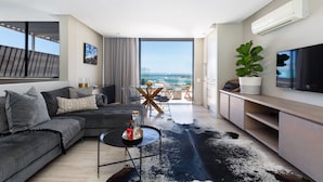 A modern living room with a large sofa, a coffee table, and a TV. There is a balcony with a view of the city. - Living Hotel Lions Eye (Cape Town)