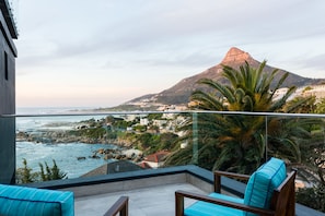 Un balcon avec deux chaises à rayures bleues, une rambarde en verre et une vue sur une ville côtière, avec ses bâtiments et une montagne en arrière-plan.