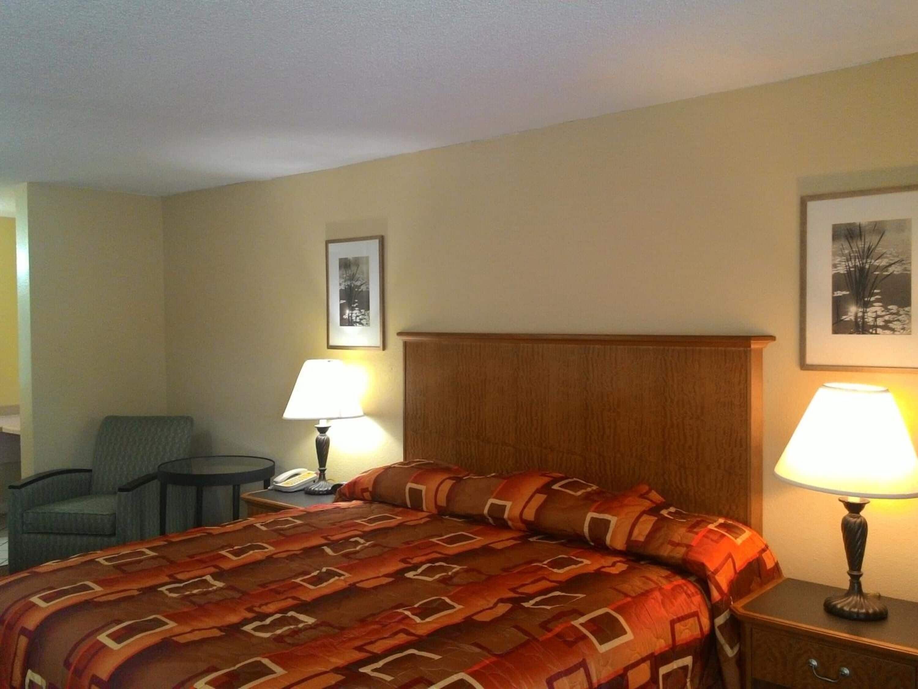 Quarto de hotel com cama, duas luminárias de cabeceira, uma cadeira, uma mesinha com telefone e duas imagens emolduradas na parede.