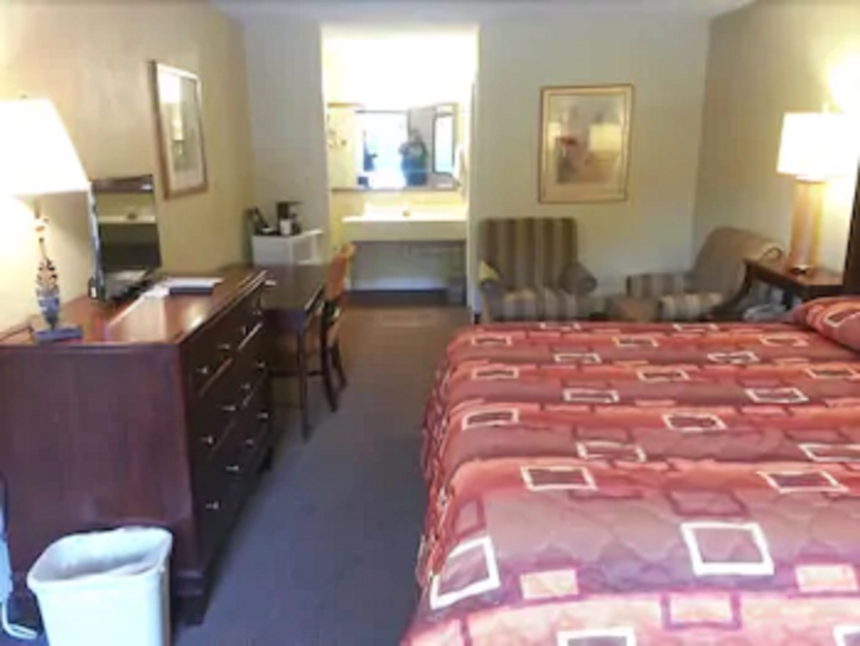 A hotel room with a bed, a desk, a chair, a dresser, a lamp, and a mirror.