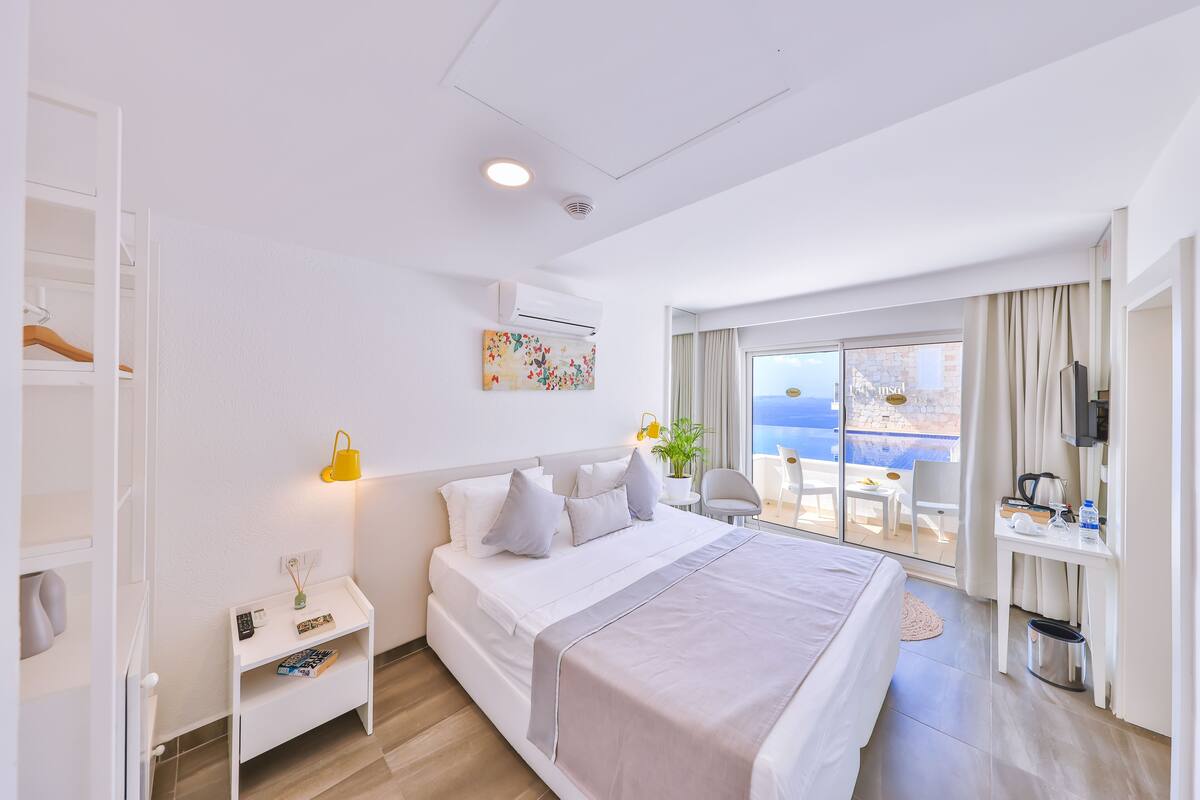 A hotel room with a bed, bedside table, and a desk with a chair. There is a window with a view of the sea.