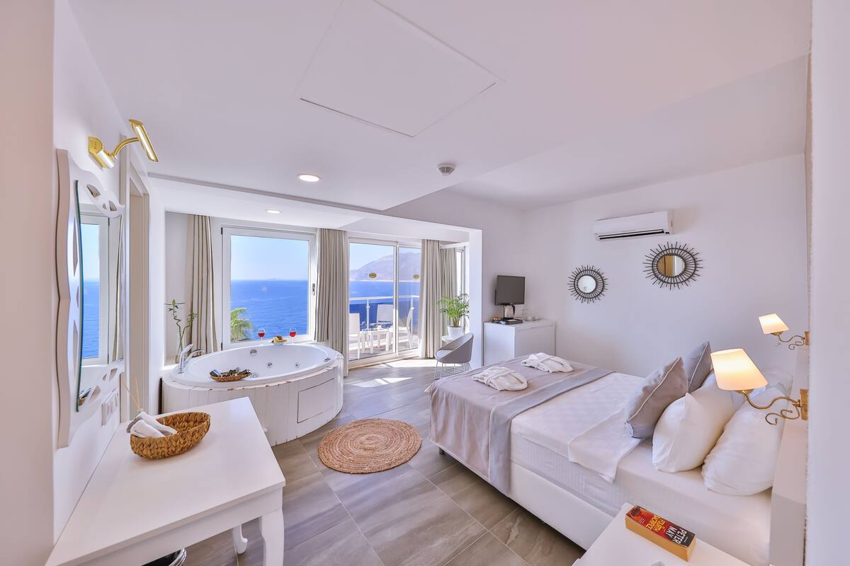 A modern hotel room with a large bathtub, a bed, a desk, and a view of the sea.