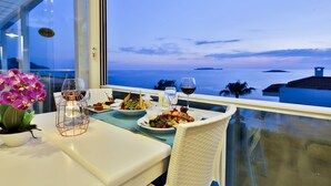 Una mesa de comedor con platos de comida, copas de vino y una vela en un balcón con vista al mar.