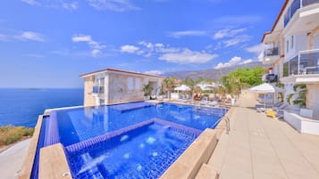 A swimming pool with a view of the sea, surrounded by lounge chairs and a building with a stone facade.
