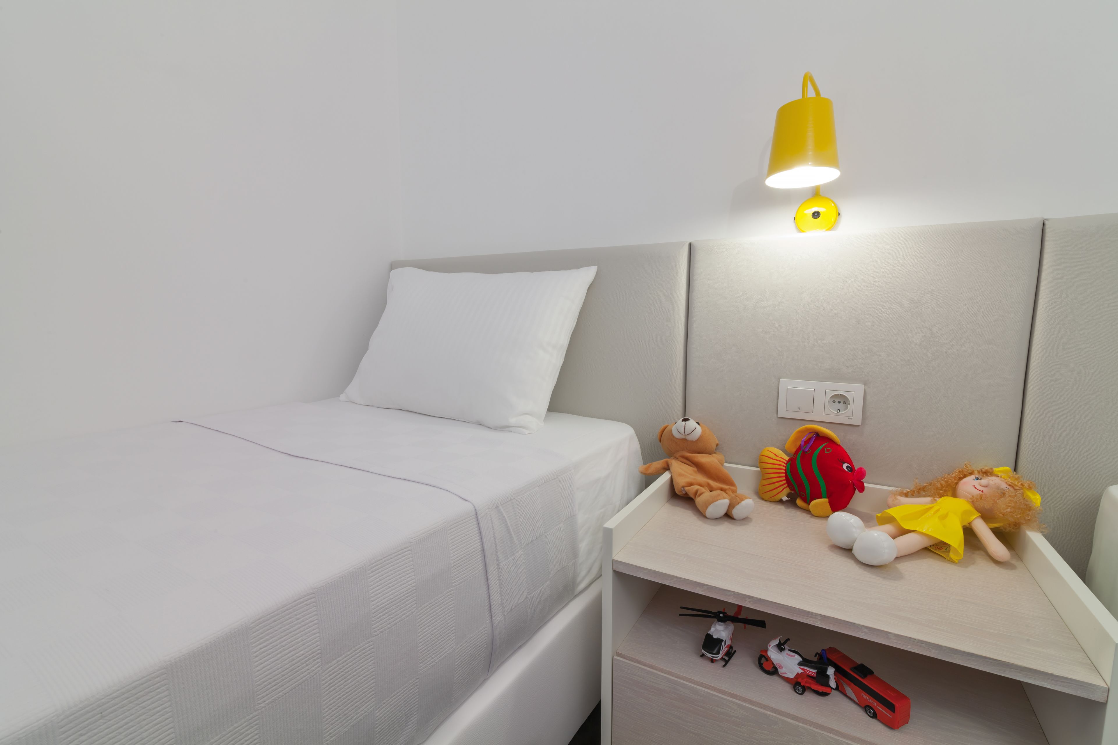 Una cama bien hecha con ropa de cama blanca, una mesita de noche con juguetes y una lámpara amarilla.