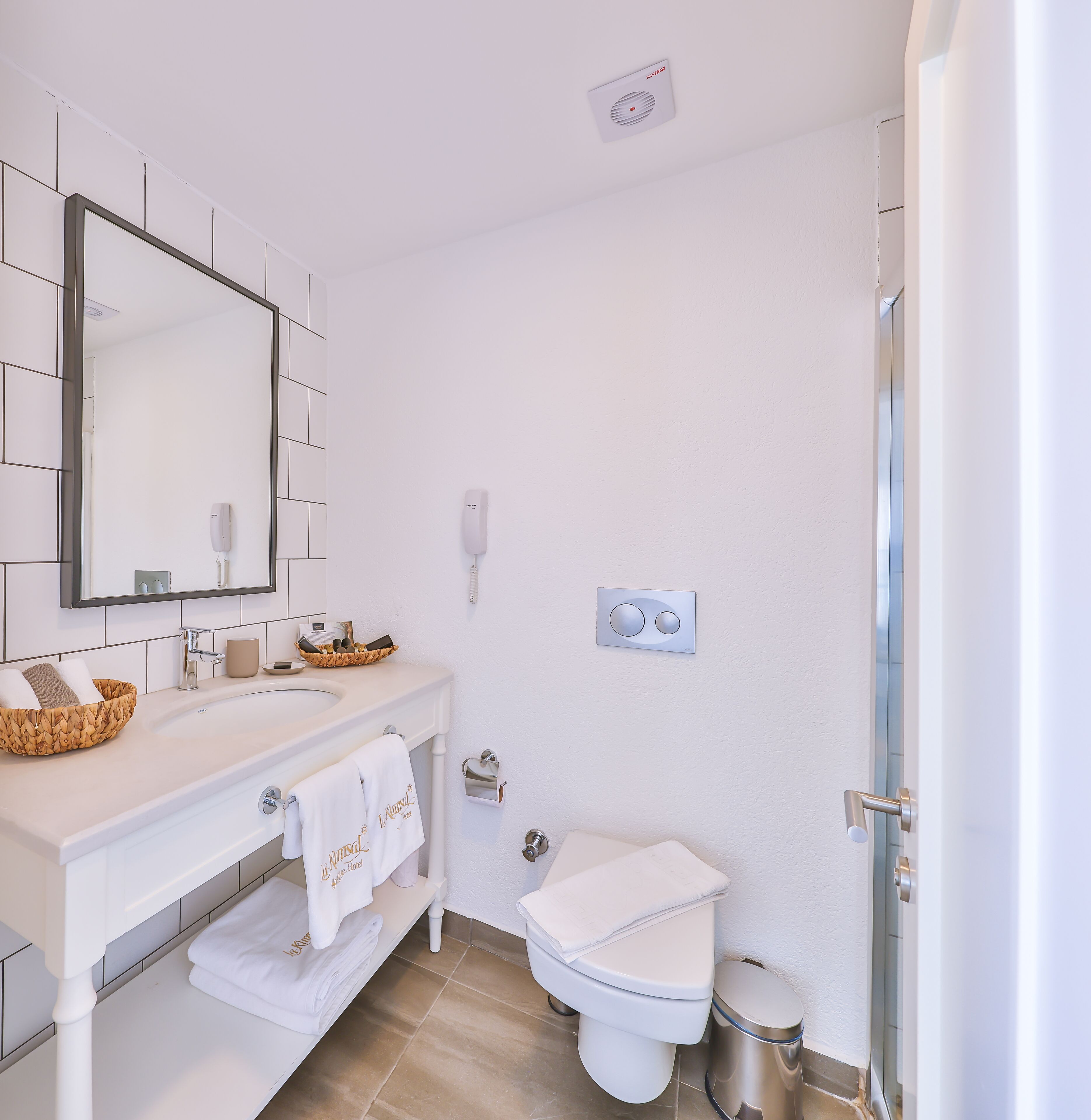 A modern bathroom with a white sink, mirror, and toilet. There are towels and toiletries on the counter, and a trash can is visible.