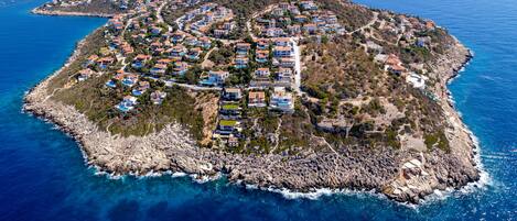 An aerial view of a coastal residential area with houses arranged in a circular pattern around a central road, surrounded by rocky terrain and the sea.
