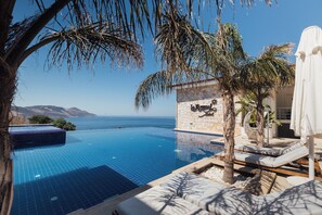 A infinity pool with a clear view of the sea, a stone wall with a sign, and lounge chairs. - La Kumsal Hotel (Kas)