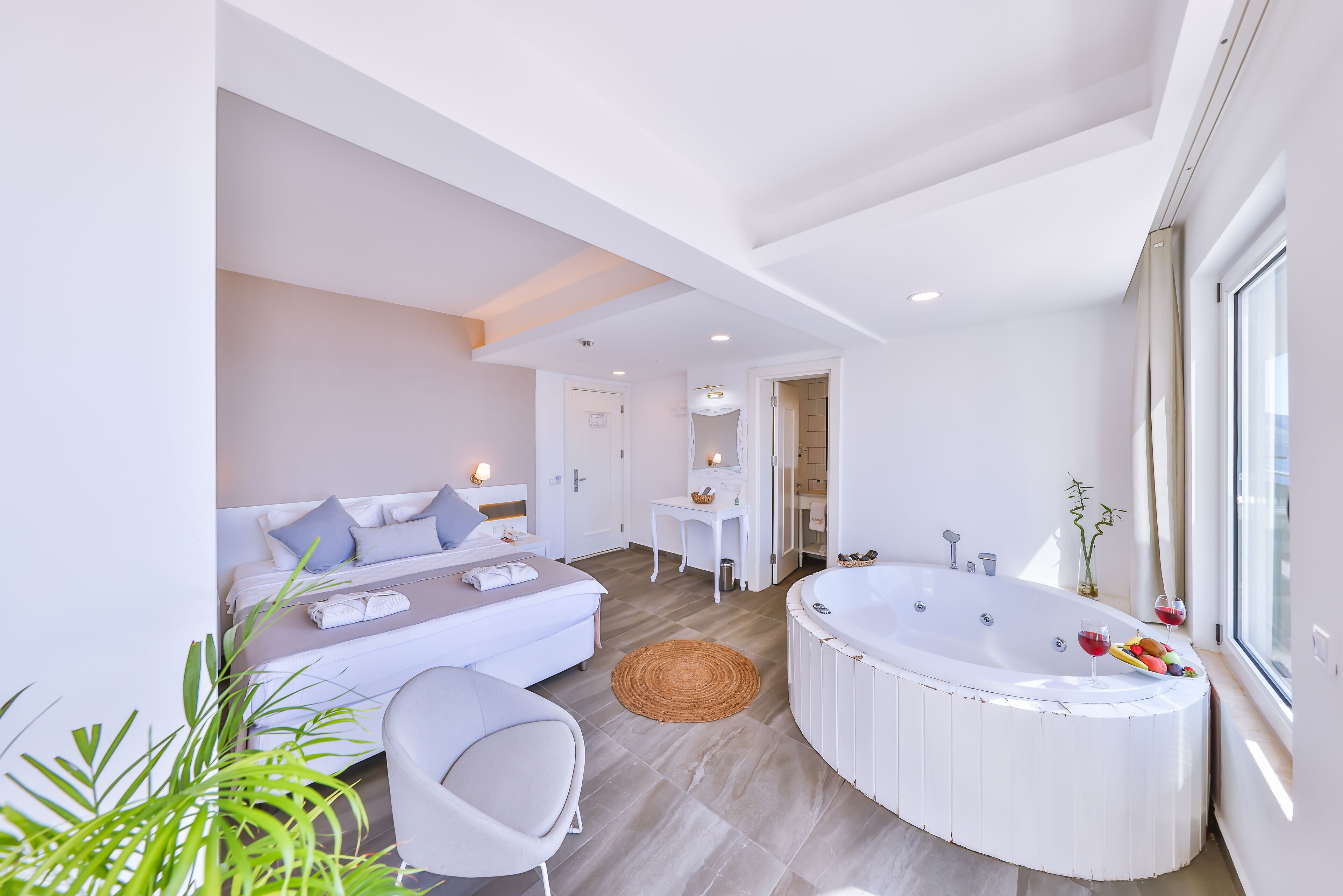 A modern bedroom with a large bed, a sitting area, a bathroom with a bathtub, and a window providing natural light.