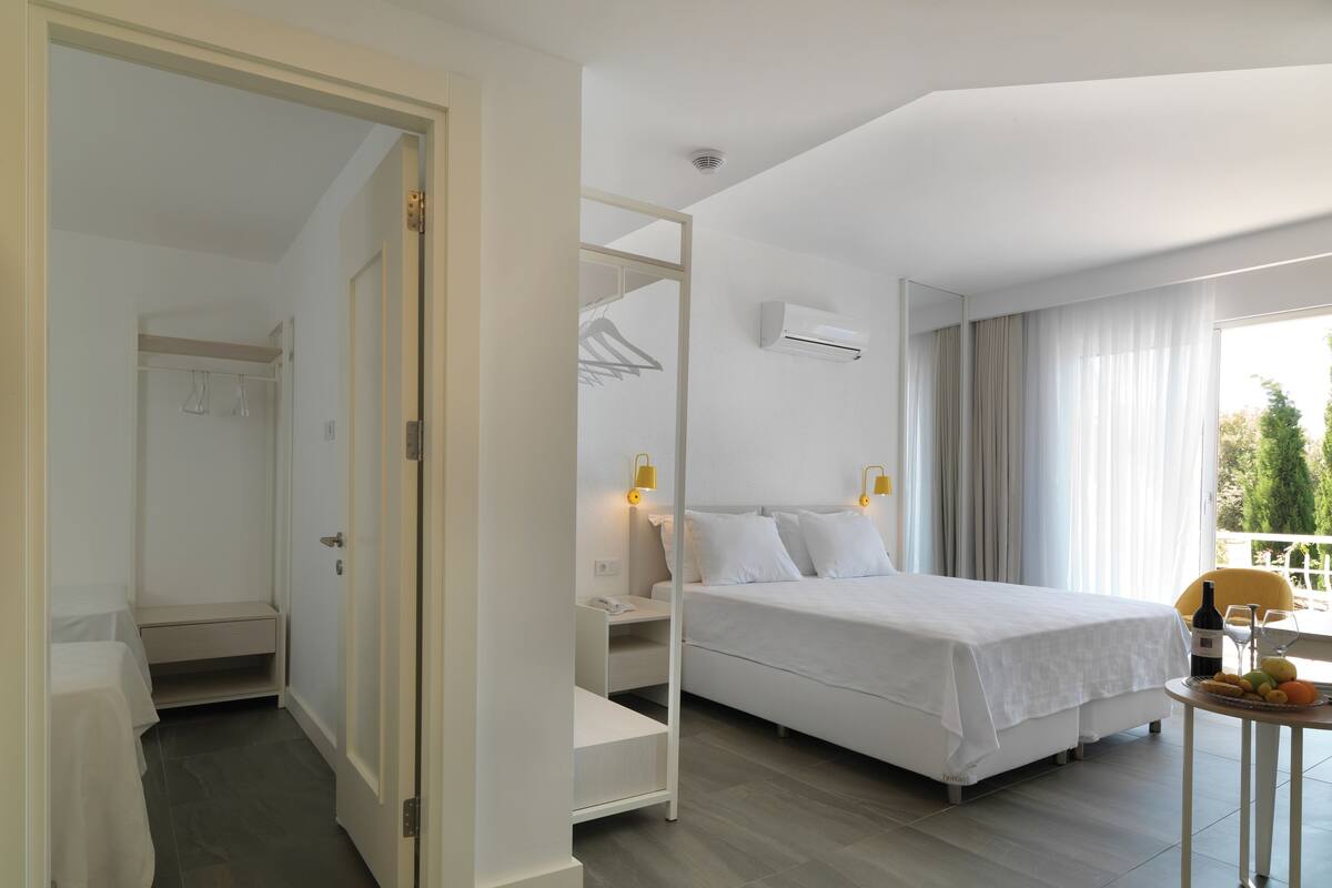 A hotel room with a bed, a small table with a bottle and glasses, a wardrobe, and a balcony with a chair.