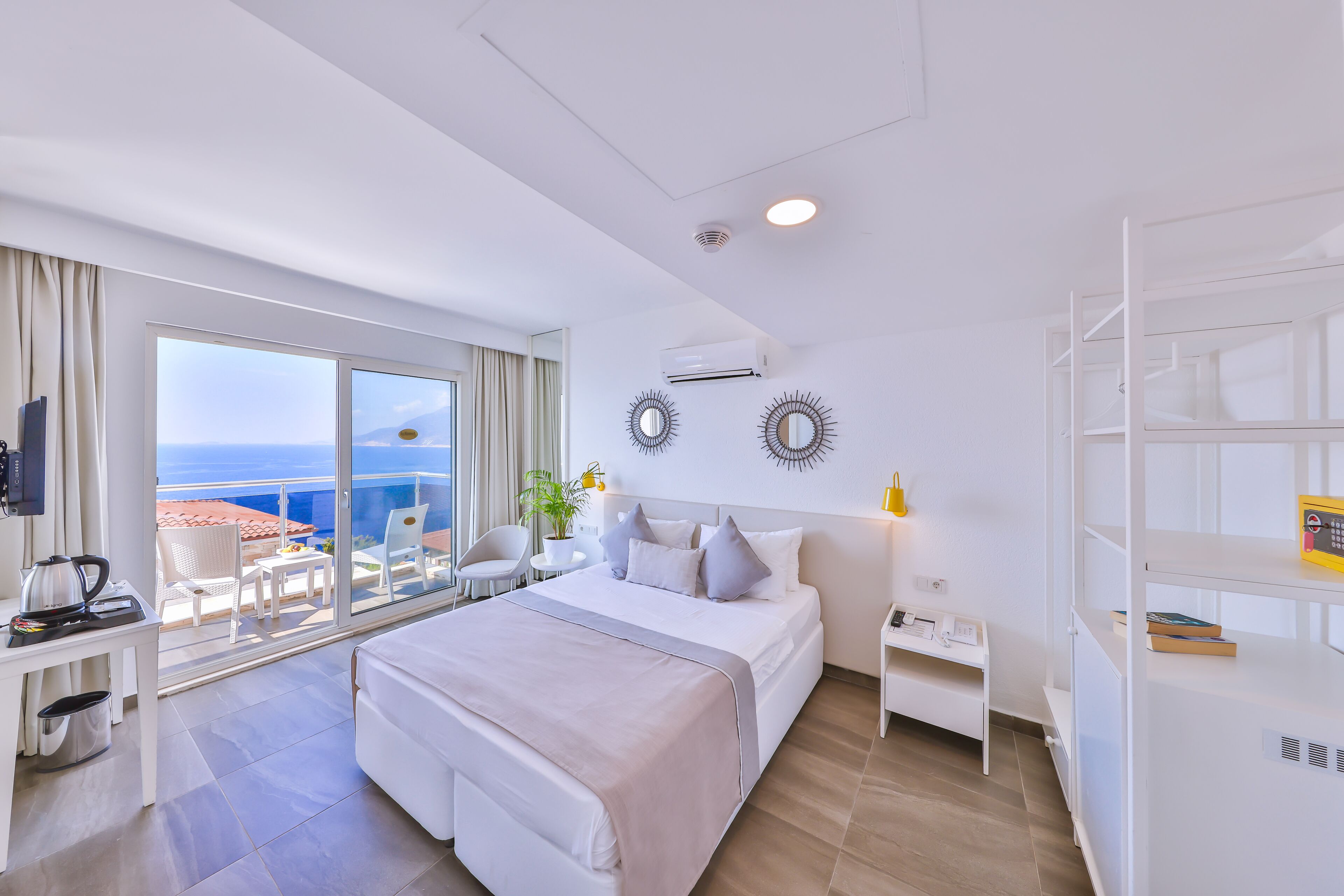A modern hotel room with a large bed, a desk with a kettle, a small table with a plant, and a balcony with a view of the sea.
