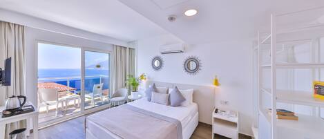 A modern hotel room with a large bed, a desk with a kettle, a small table with a plant, and a balcony with a view of the sea.