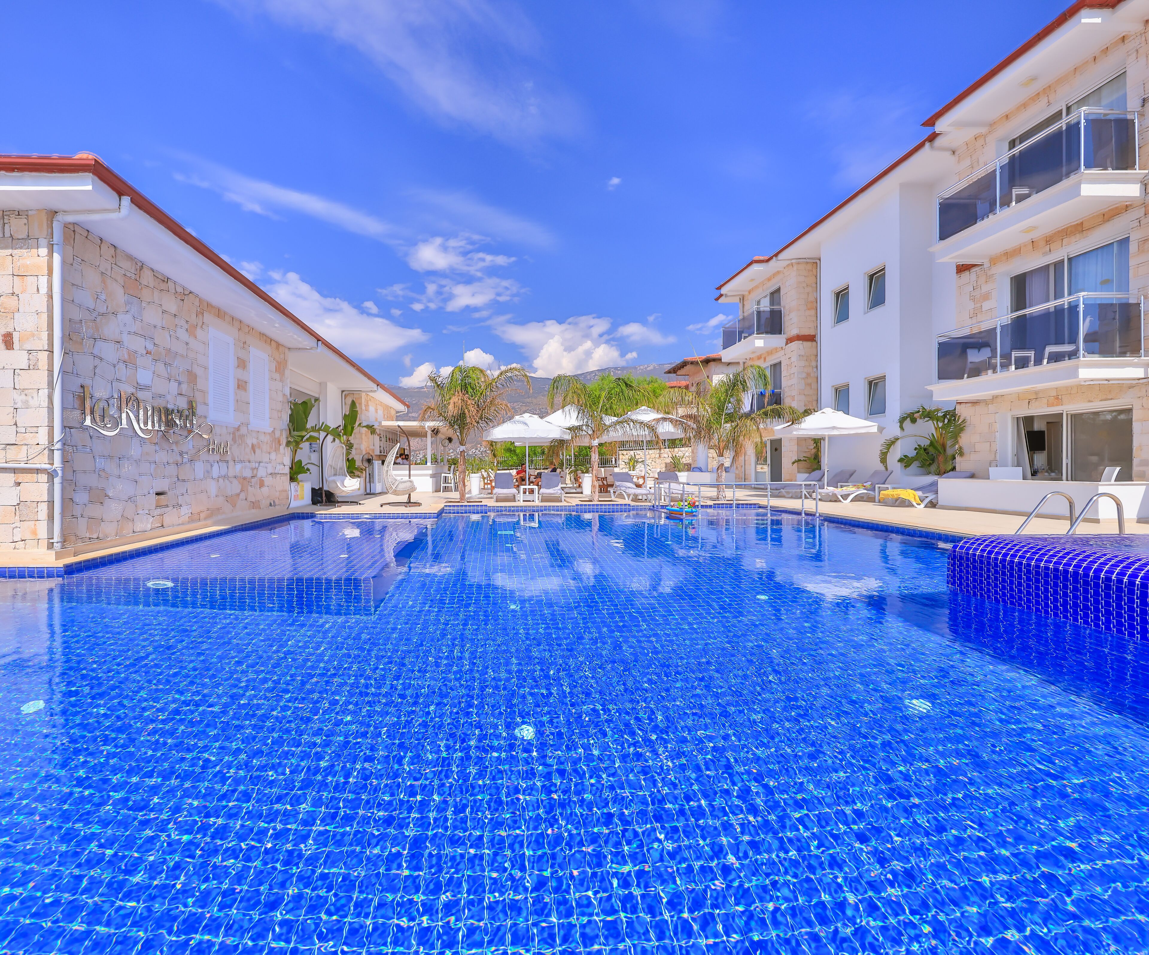 A swimming pool with tiled blue surface, surrounded by lounge chairs and umbrellas, and adjacent buildings with balconies.