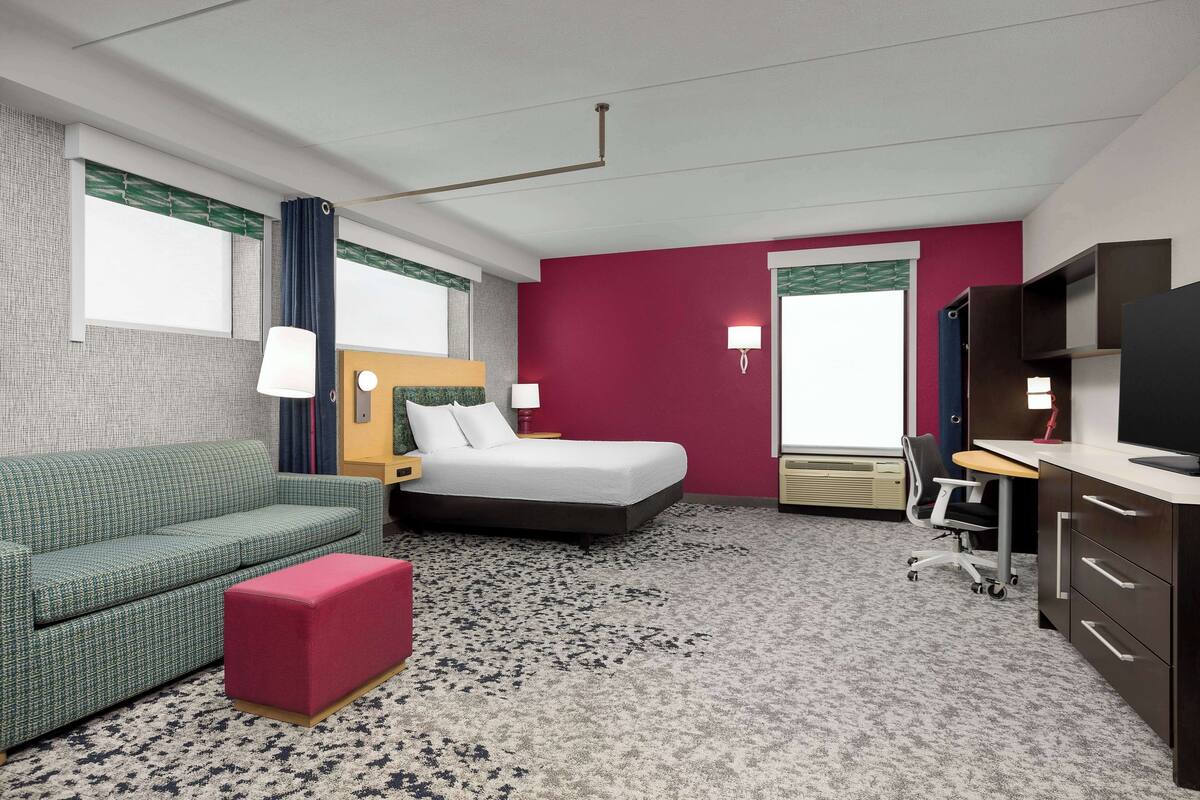 Une chambre d’hôtel avec un canapé à motifs verts, un pouf rouge, un lit avec du linge de lit blanc, un bureau avec une chaise et un téléviseur posé sur un meuble.